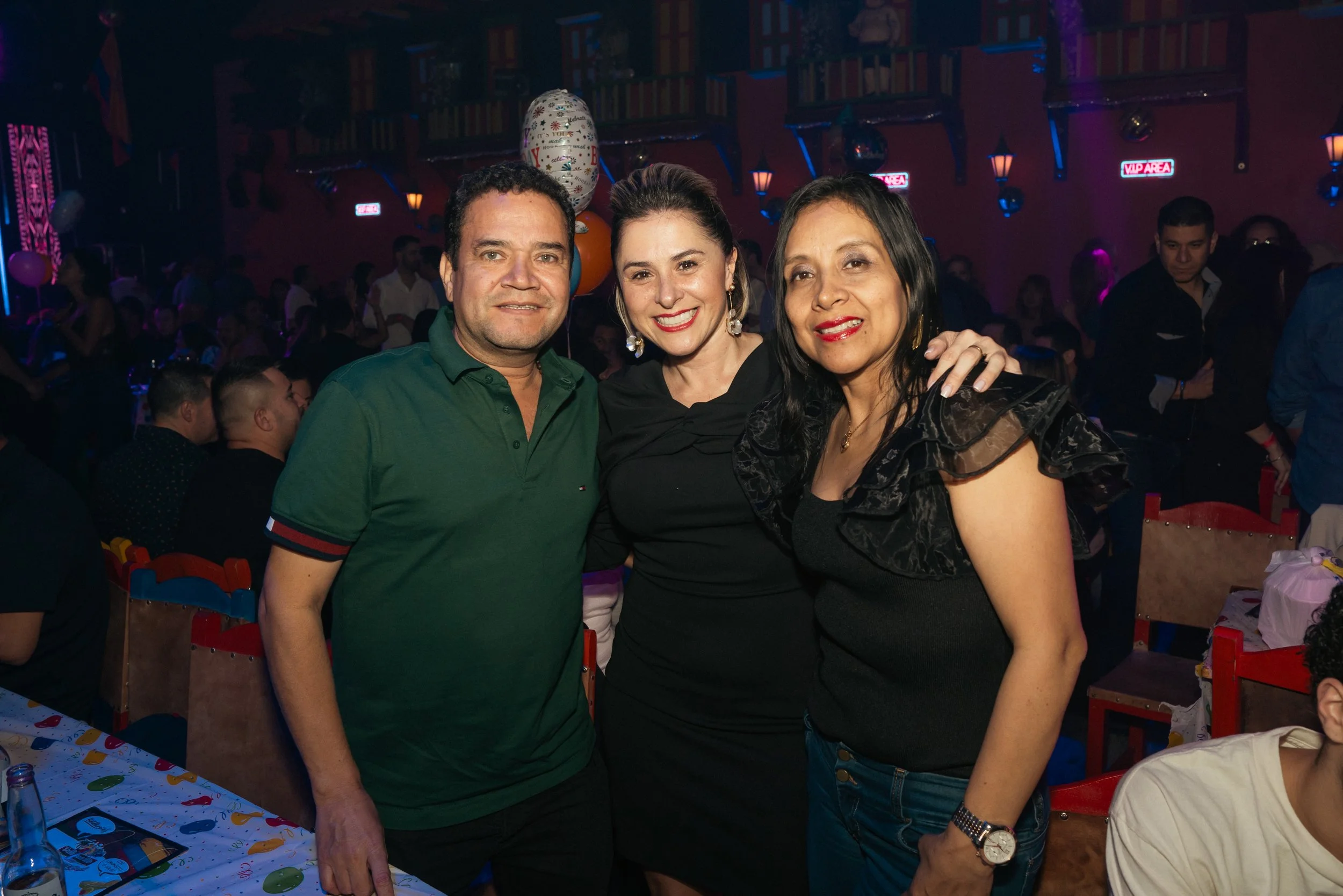 Three people smiling at a party or celebration in a dimly lit venue with colorful lighting and a crowd in the background.