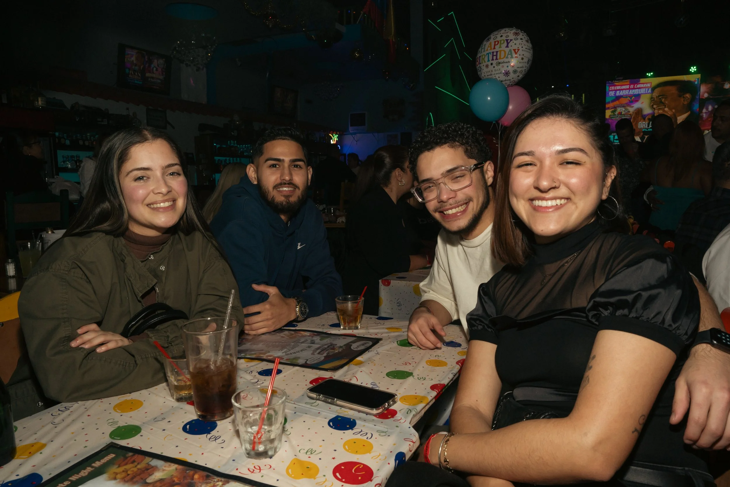 Group of four friends smiling while dining at a restaurant decorated for a birthday celebration with balloons including one that says 'Happy Birthday'.