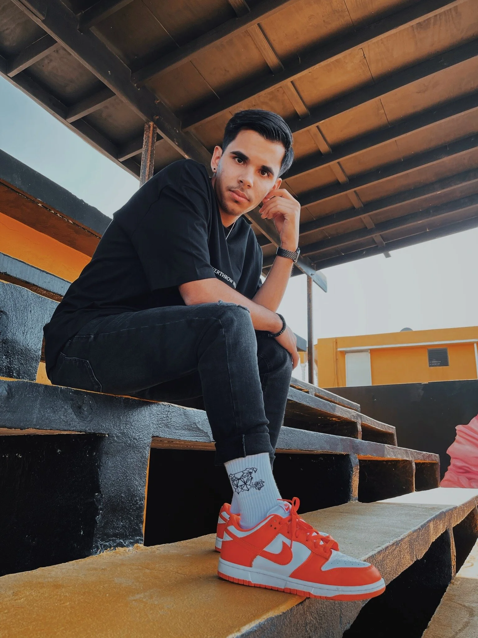 A young man sitting on outdoor stairs under a wooden roof, wearing a black t-shirt, black jeans, white socks with a black design, and red and white sneakers.