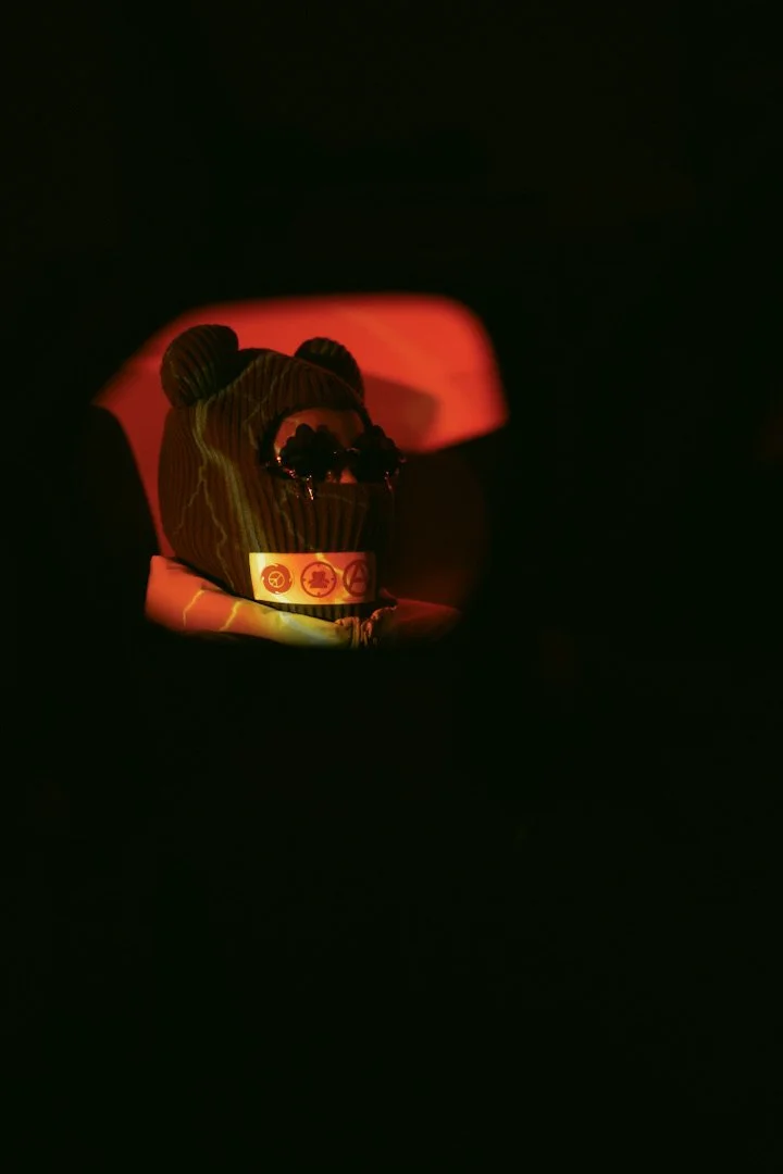 A stuffed animal wearing sunglasses and a band with symbols, illuminated by a red light in a dark environment.