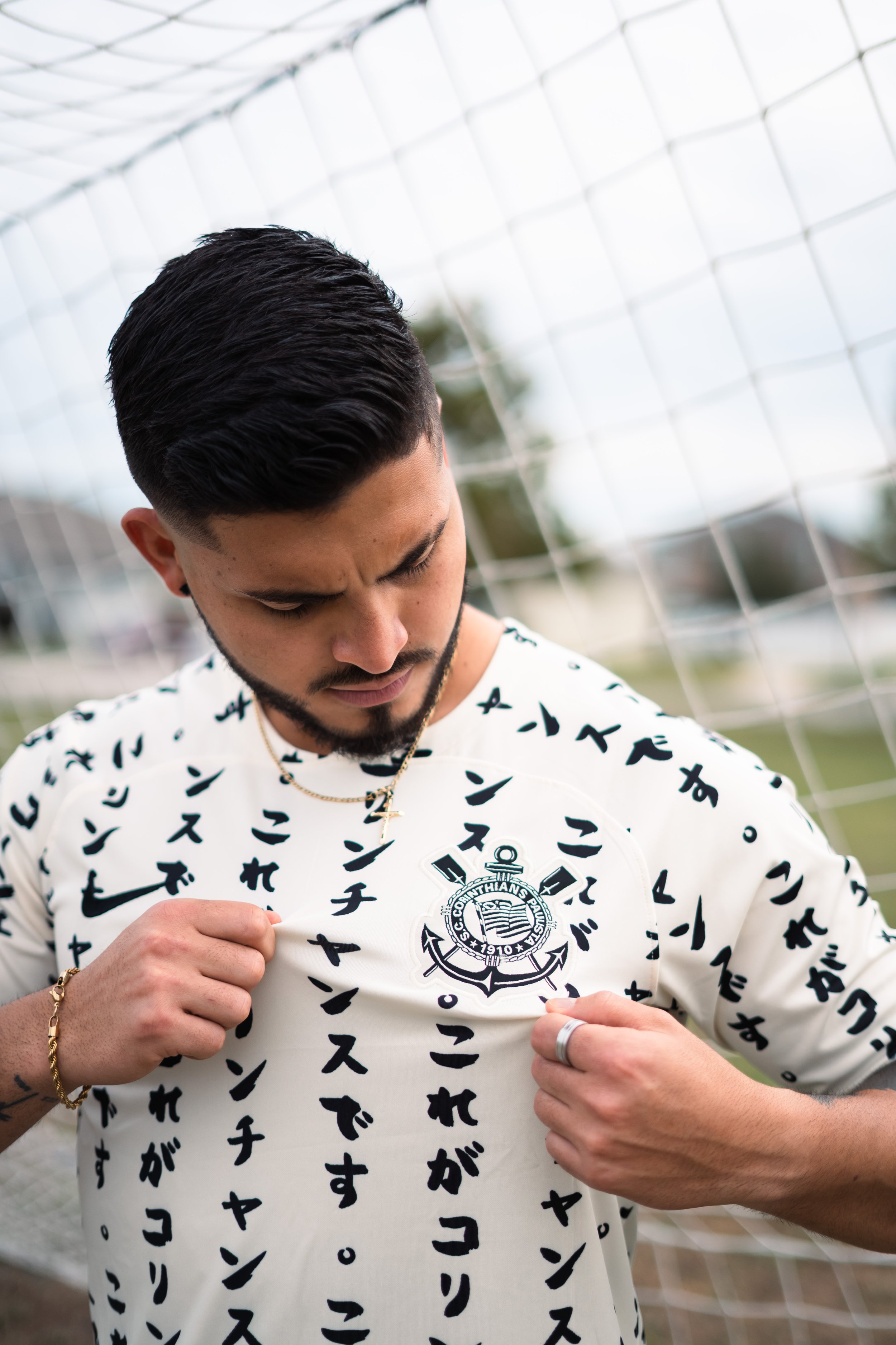 Man with dark hair and beard wearing a white soccer jersey with black Japanese characters, looking down and pulling up the jersey to reveal a Corinthians badge on his chest. He is standing outdoors near a goal net.