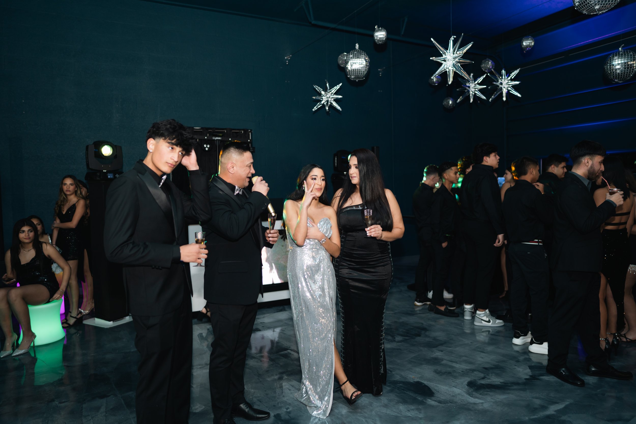 People at a formal party standing and sitting, with some talking into a microphone, wearing elegant black and silver dresses and suits, with holiday decorations hanging from the ceiling.