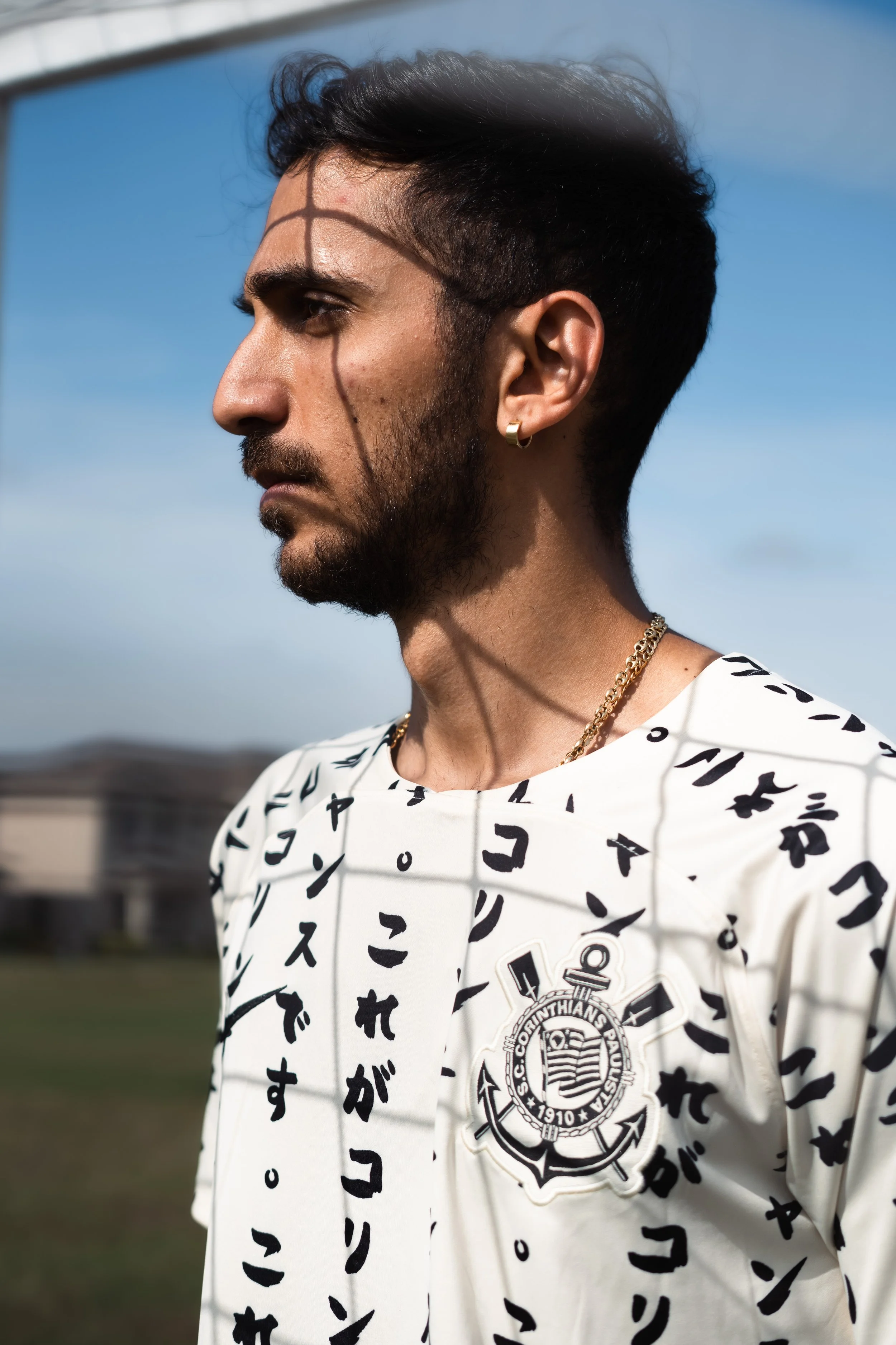 A man with dark hair and beard wearing a white sports jersey with Japanese characters and the Corinthians football club insignia, standing outdoors on a sunny day.
