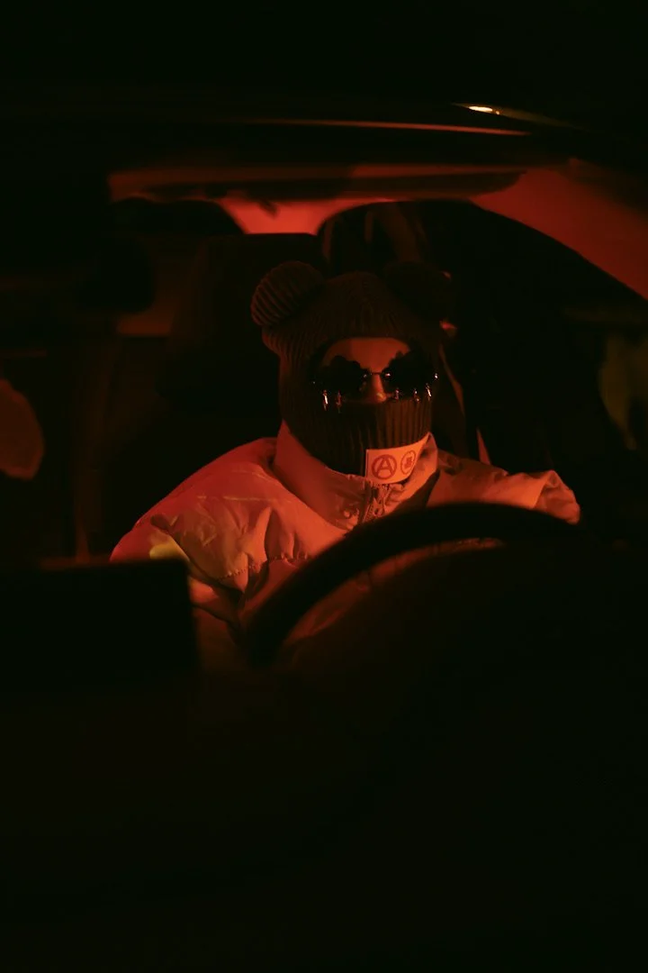 Person sitting in the driver's seat of a car at night, wearing a beanie, sunglasses, and a jacket, with a dark and red-colored lighting ambiance.