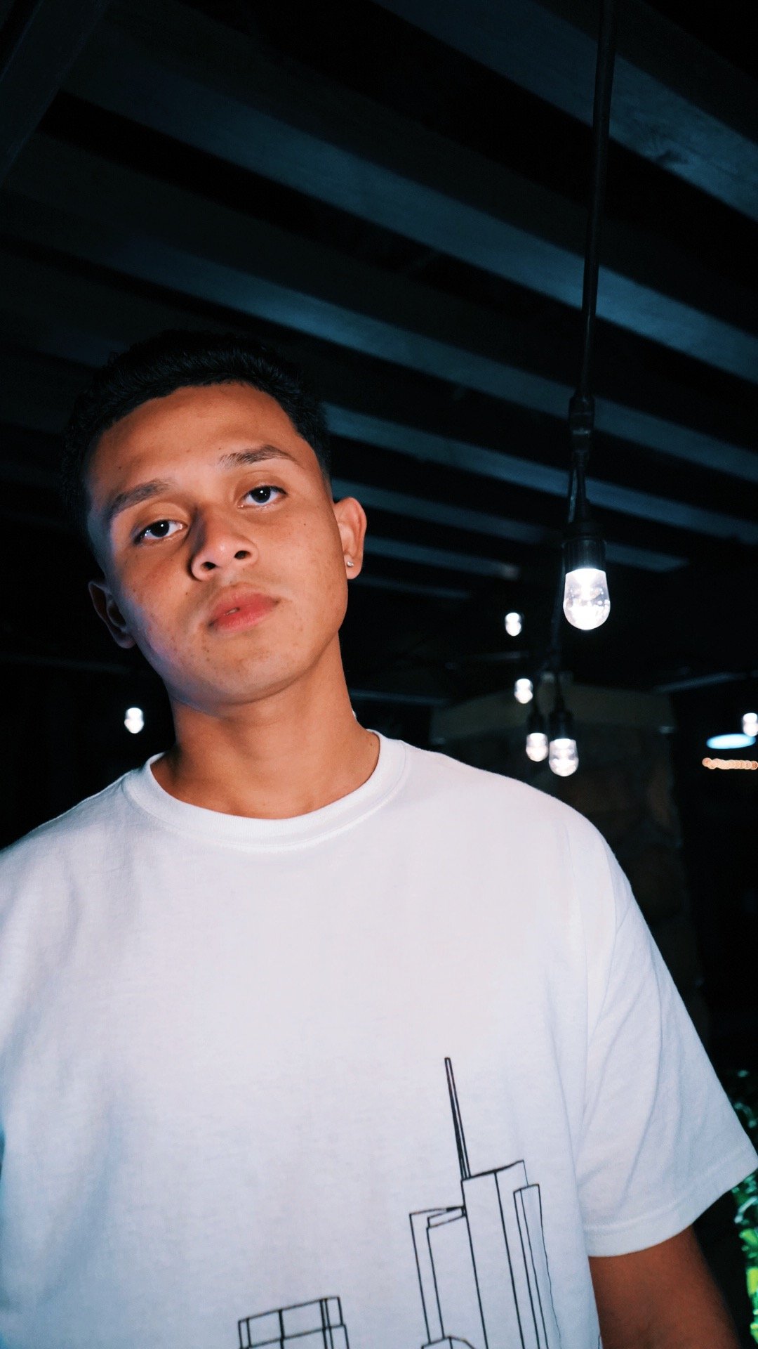 A young man with short dark hair and small earrings wearing a white t-shirt with a minimalist line art design, standing indoors with dim lighting and hanging light bulbs in the background.