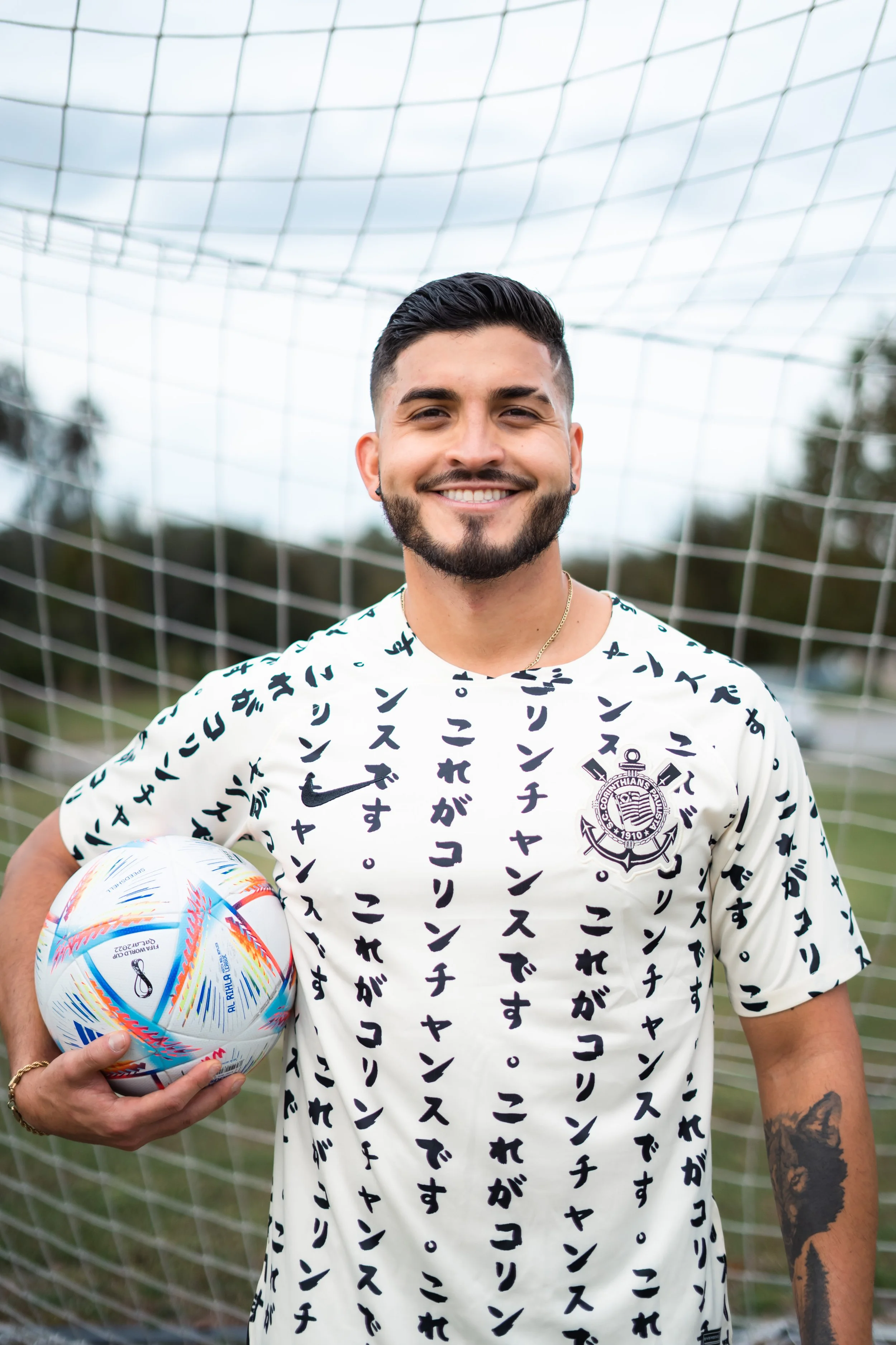 A young man with dark hair, a beard, and a tattoo on his left arm, wearing a white sports jersey with Japanese characters and holding a soccer ball, standing in front of a soccer net.