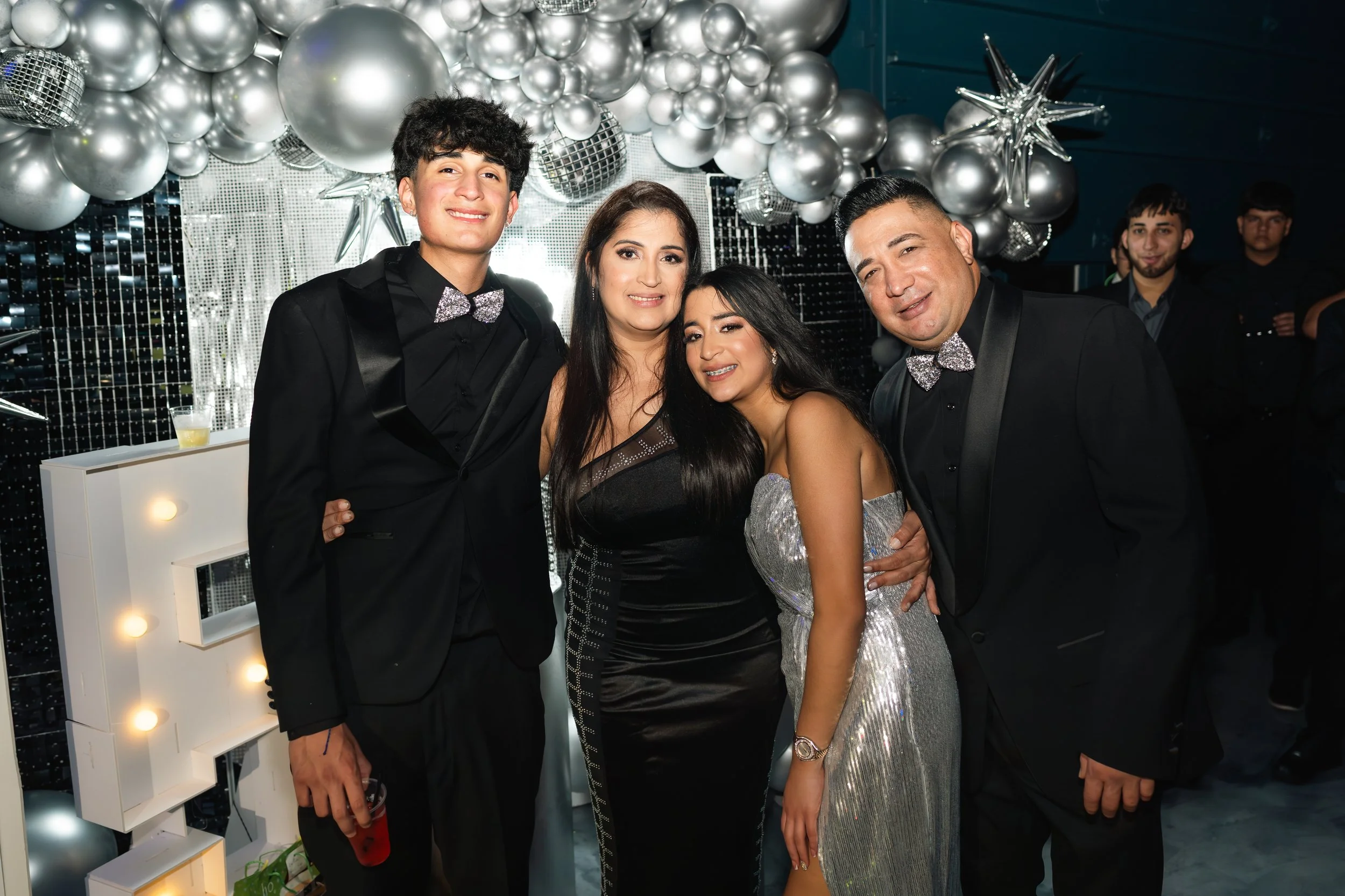 Group of four people dressed in formal attire, standing closely together at a celebration, with silver balloons, stars, and a reflective backdrop behind them.