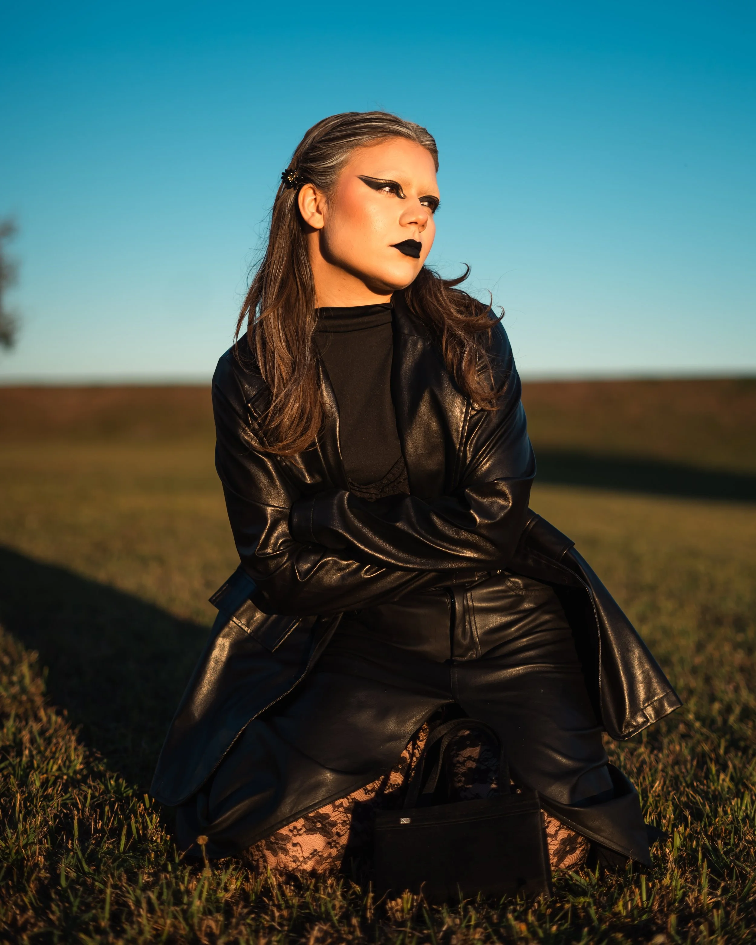 A young woman with dramatic black eyeliner and lipstick, dressed in black leather clothing, sitting on the grass outdoors during sunset.
