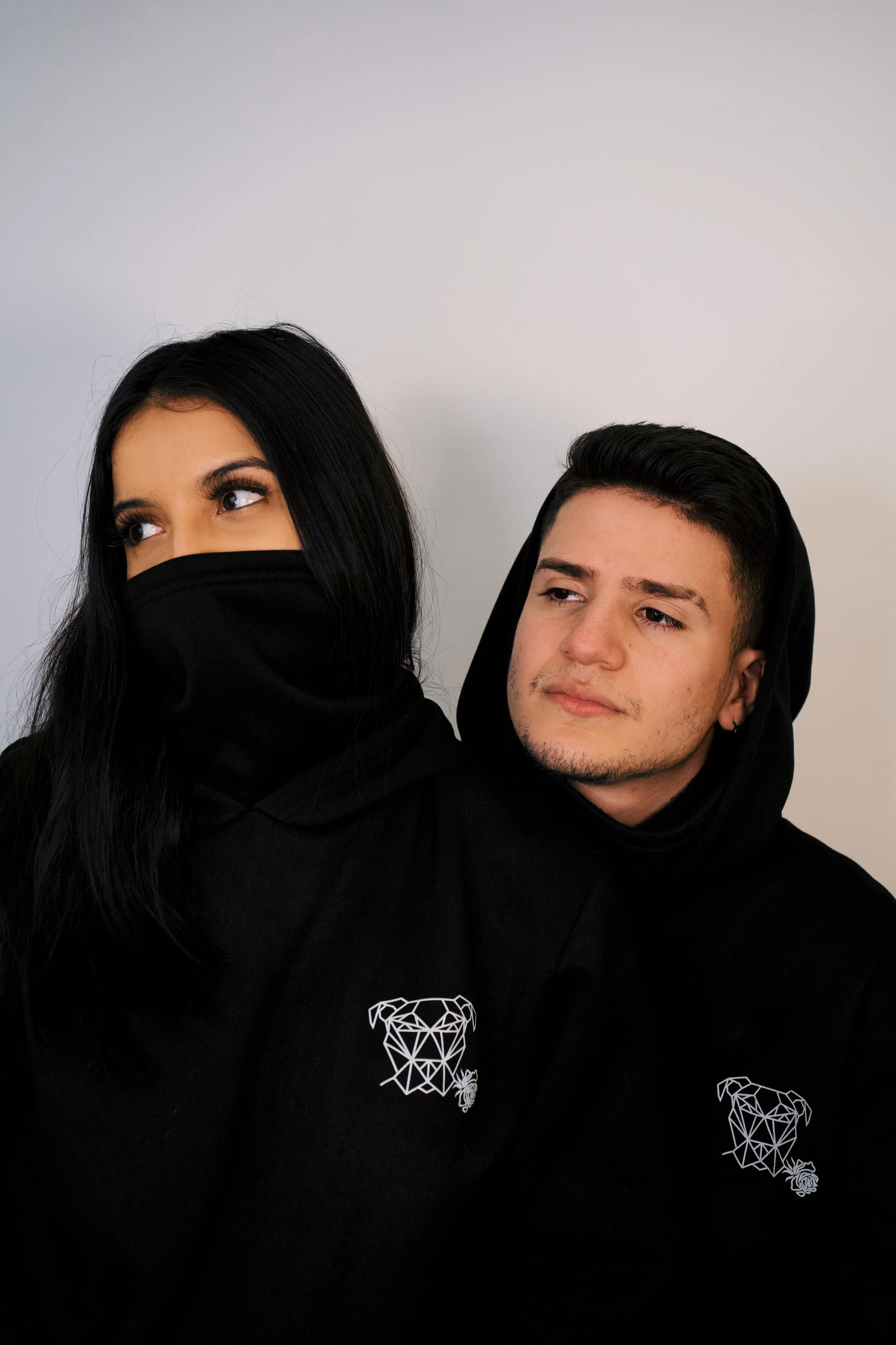 A young woman with black hair and makeup, partially covered with a black face covering, standing next to a young man with dark hair and a slight beard, both wearing black hoodies with geometric dog designs, against a plain white background.