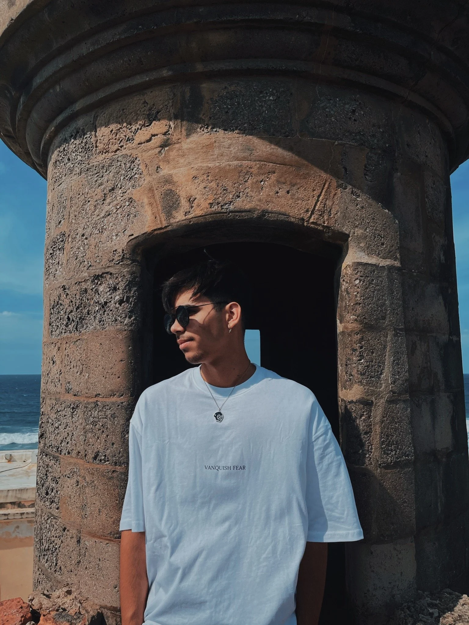 A young man wearing sunglasses and a white T-shirt with the words 'VANQUISH FEAR' printed on it, standing in front of a stone structure near the beach on a sunny day.