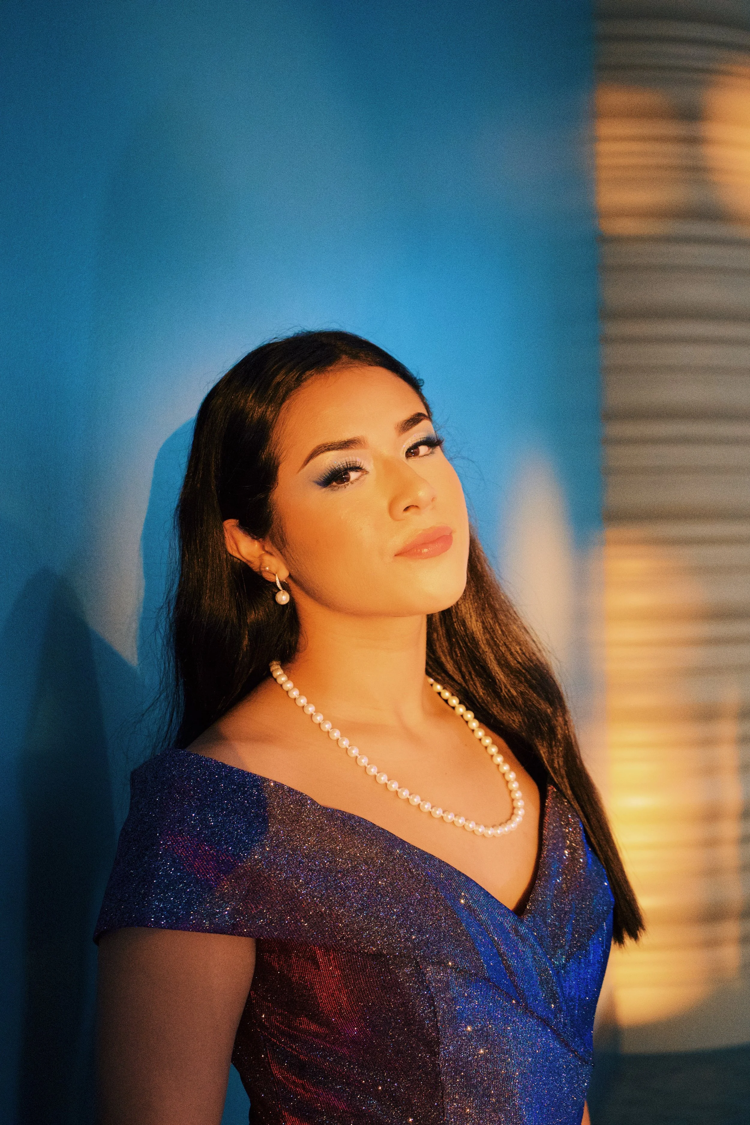 A woman with long dark hair, wearing pearl earrings, a pearl necklace, and a sparkly blue dress, standing against a blue wall with warm lighting on her face.