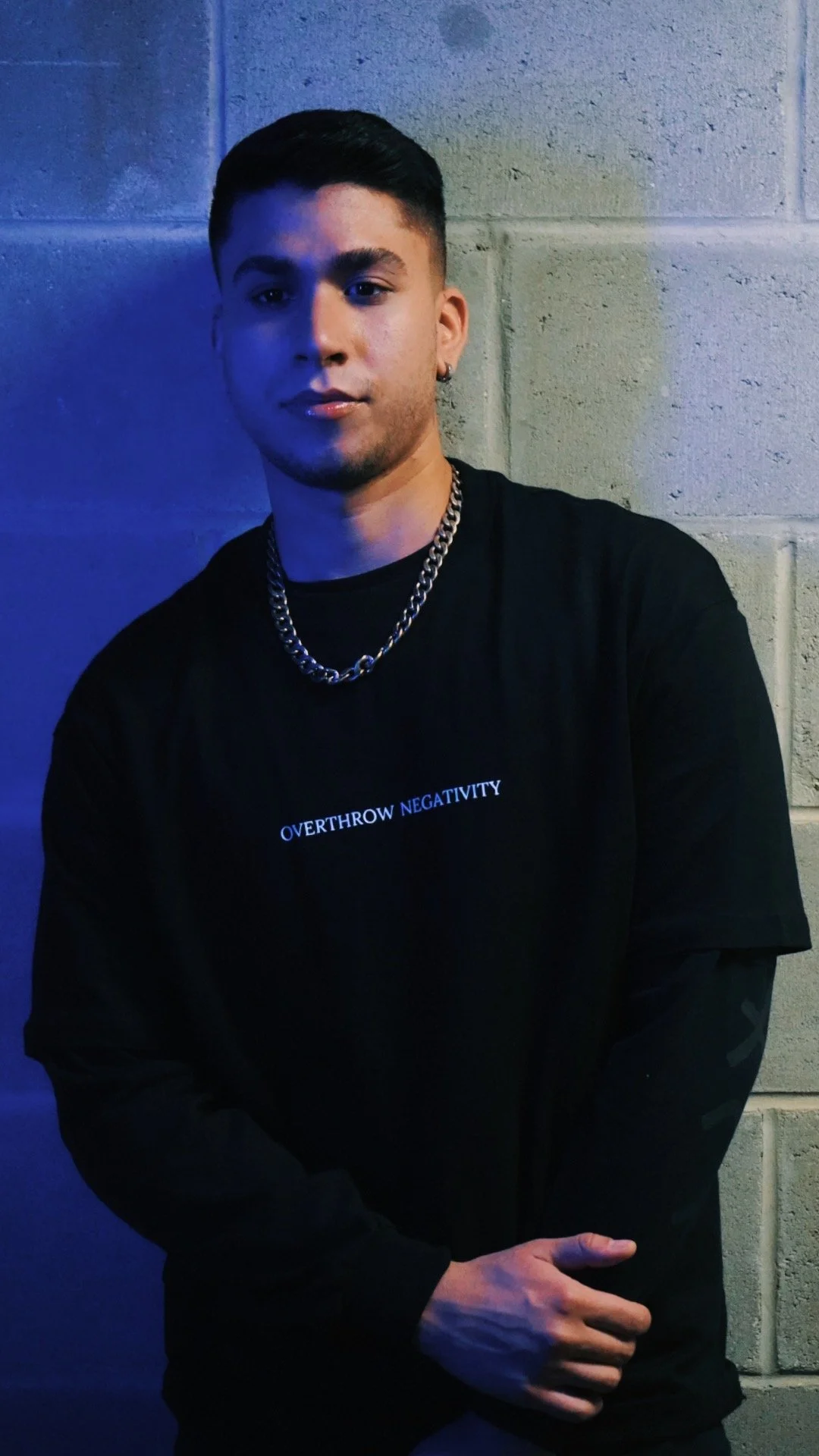 A young man with short dark hair, wearing a black T-shirt with white text, a silver chain, and a small earring, standing against a gray brick wall with colorful lighting.