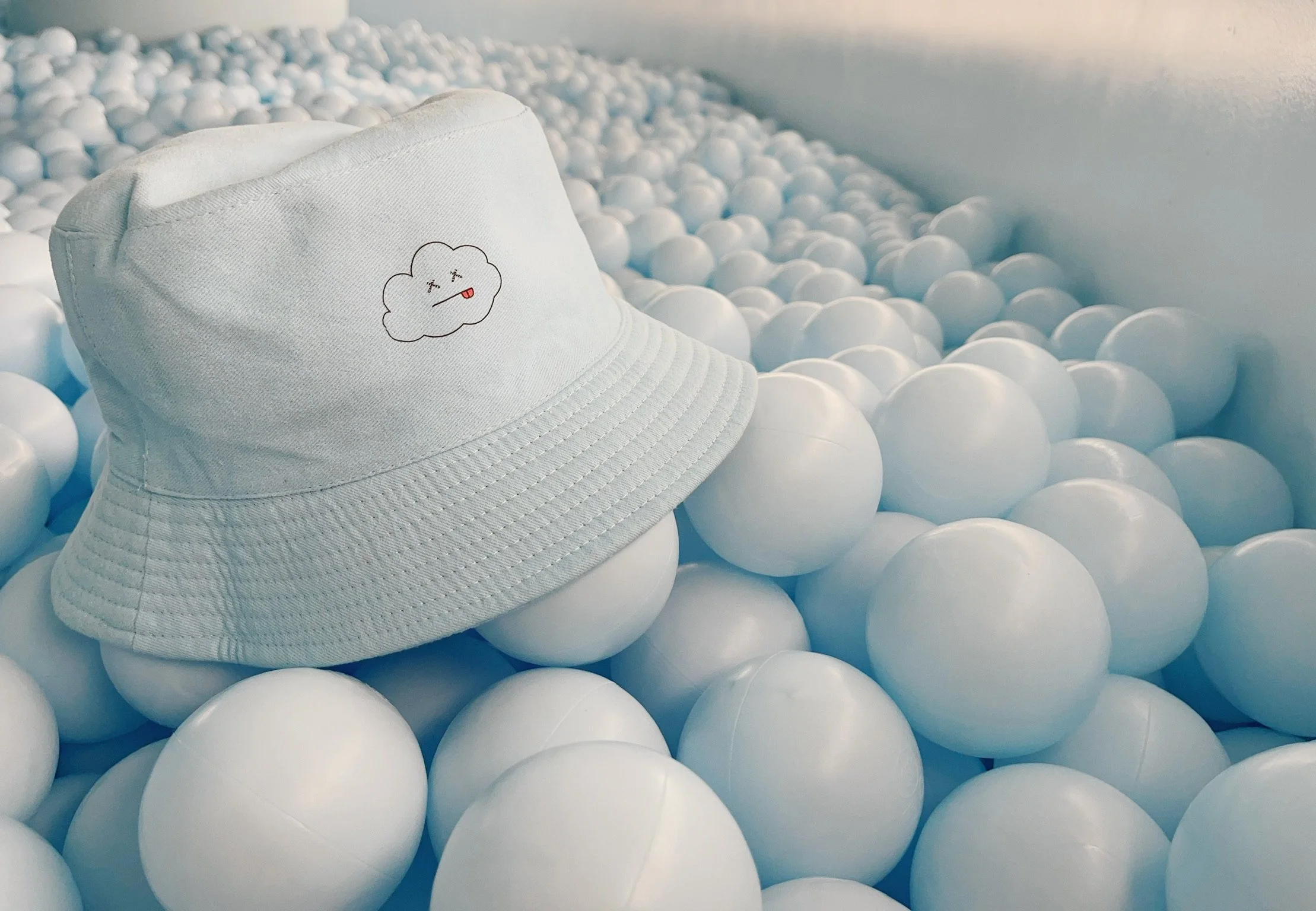 A white bucket hat with a small cartoon cloud face embroidered on it, resting among many white plastic balls in a ball pit.