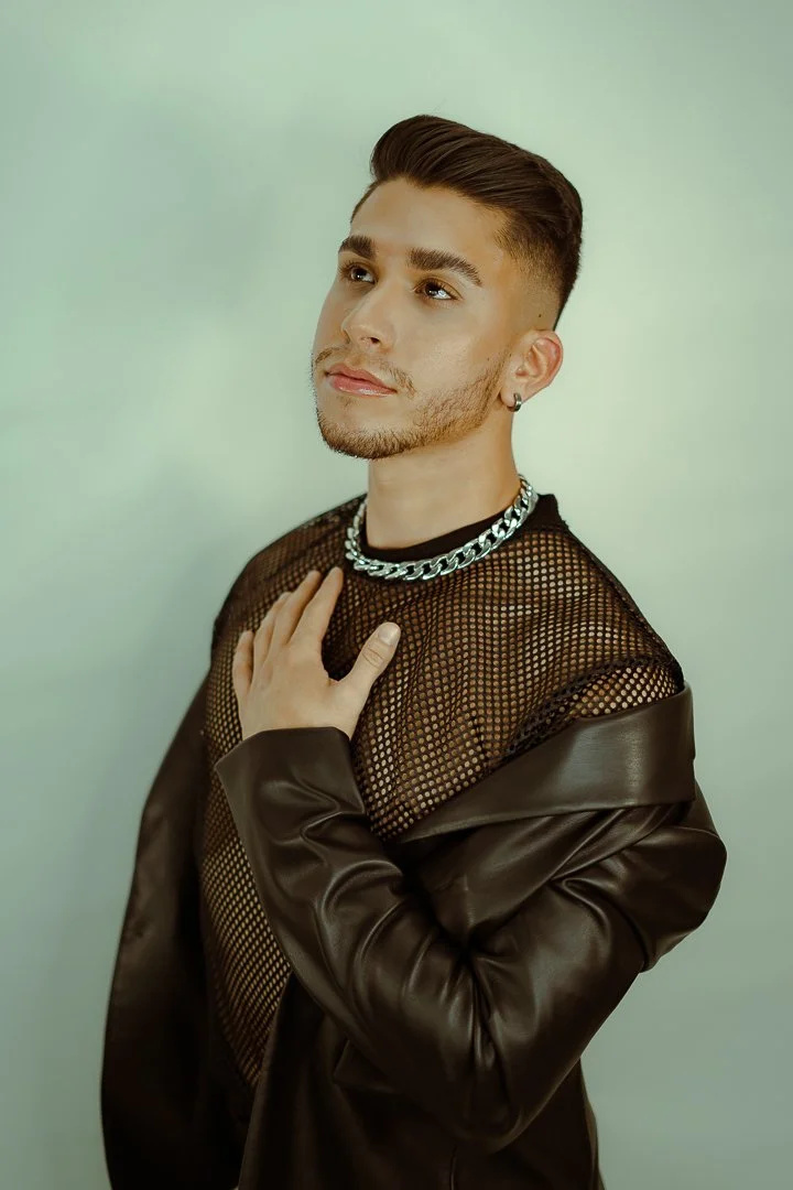 A young man with styled hair and a beard, wearing a black mesh top, a chunky silver chain necklace, and a brown leather jacket. He has one hand on his chest and gazes thoughtfully upward.