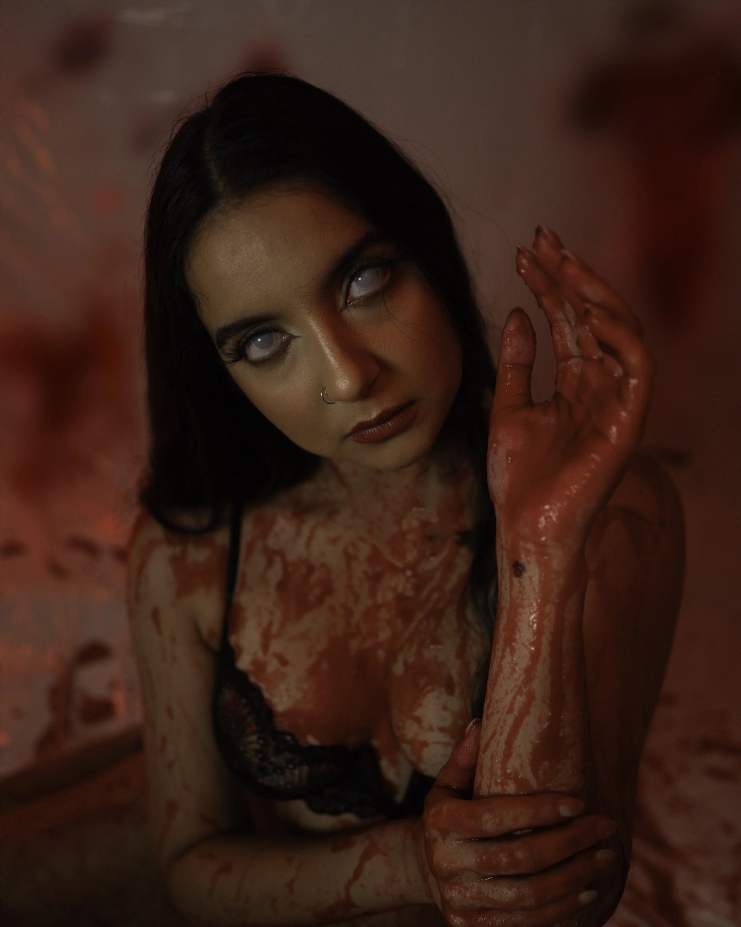 A woman with dark hair and makeup covered in fake blood, with white eyes, looking upward, with her hand covered in blood near her face.