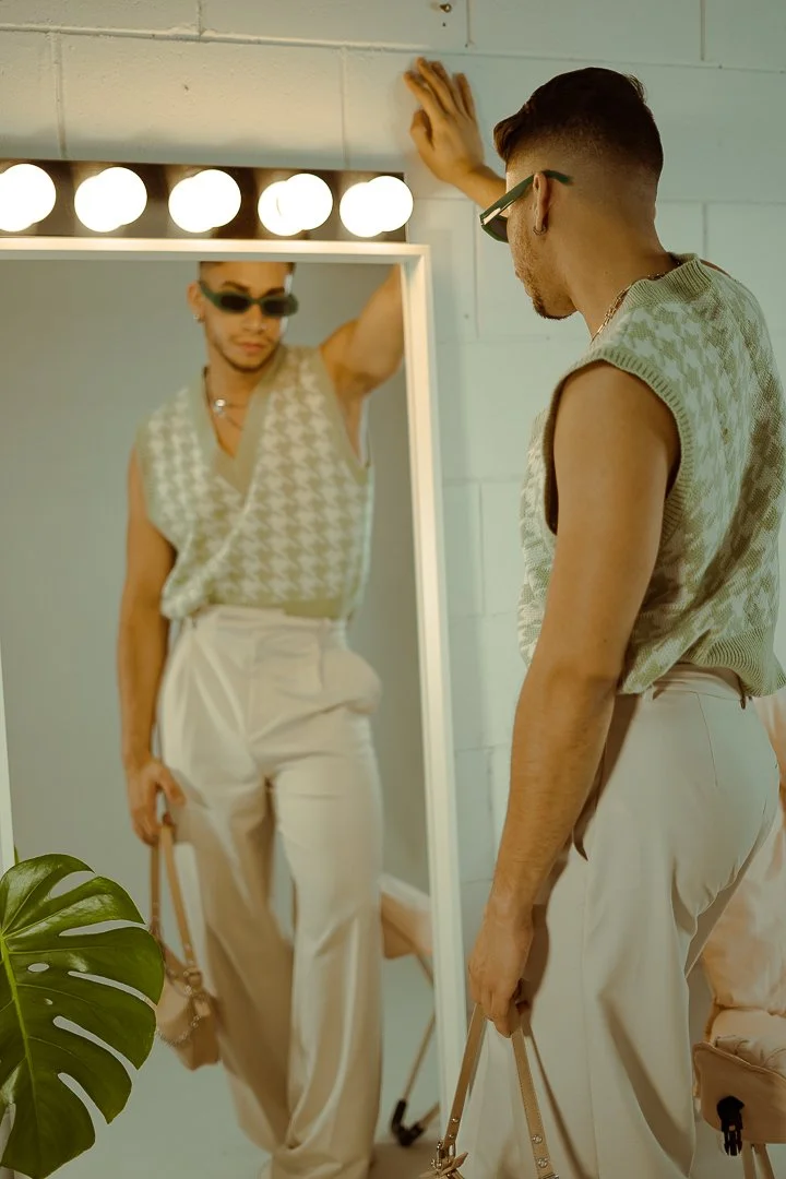 A person looking at their reflection in a mirror, wearing sunglasses, a vest, and high-waisted pants, in a room with a bright vanity light and a green plant in the foreground.