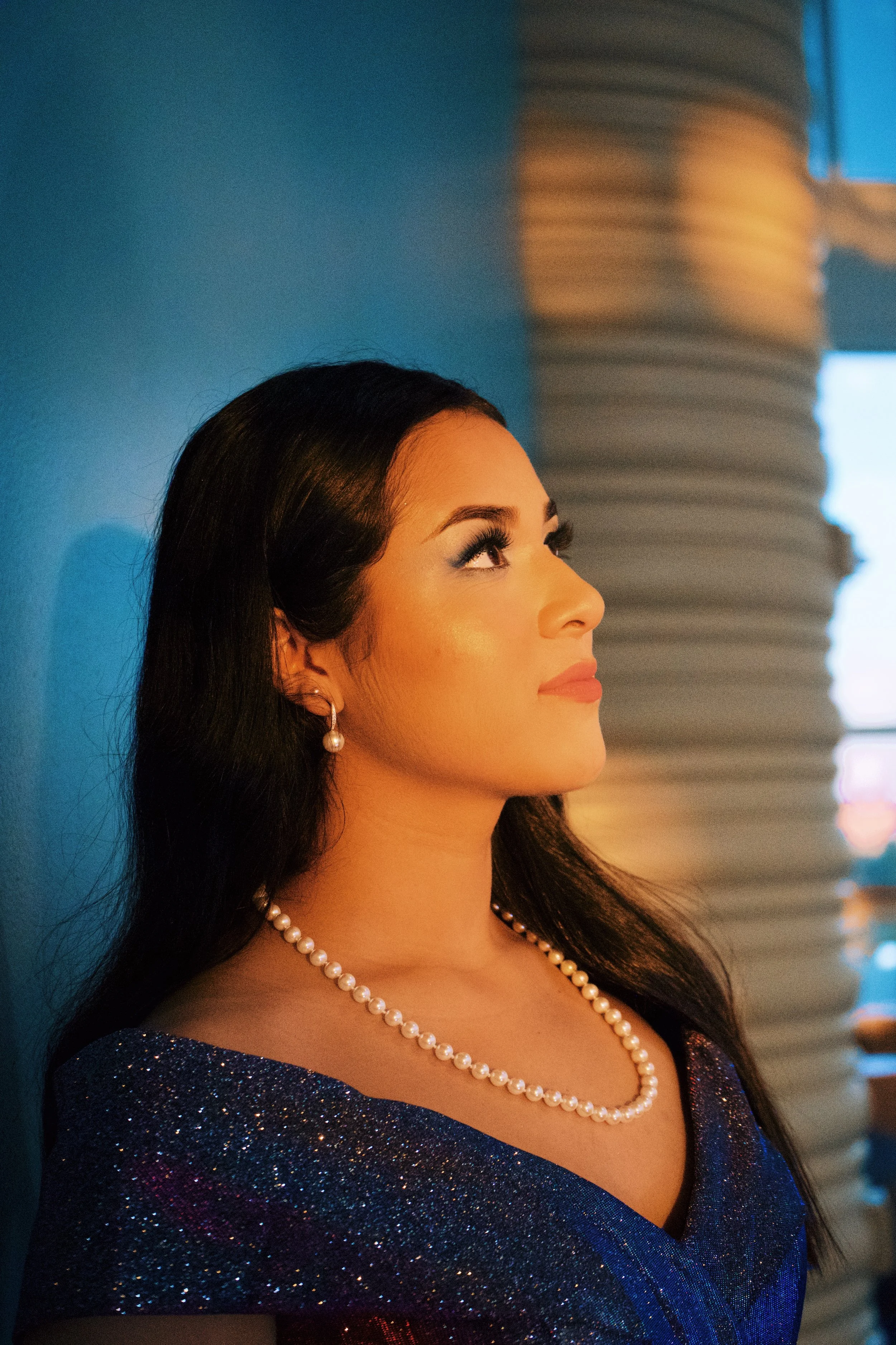 Portrait of a woman wearing pearl earrings and necklace, with long dark hair, standing against a blue background, looking to the right with a serene expression.