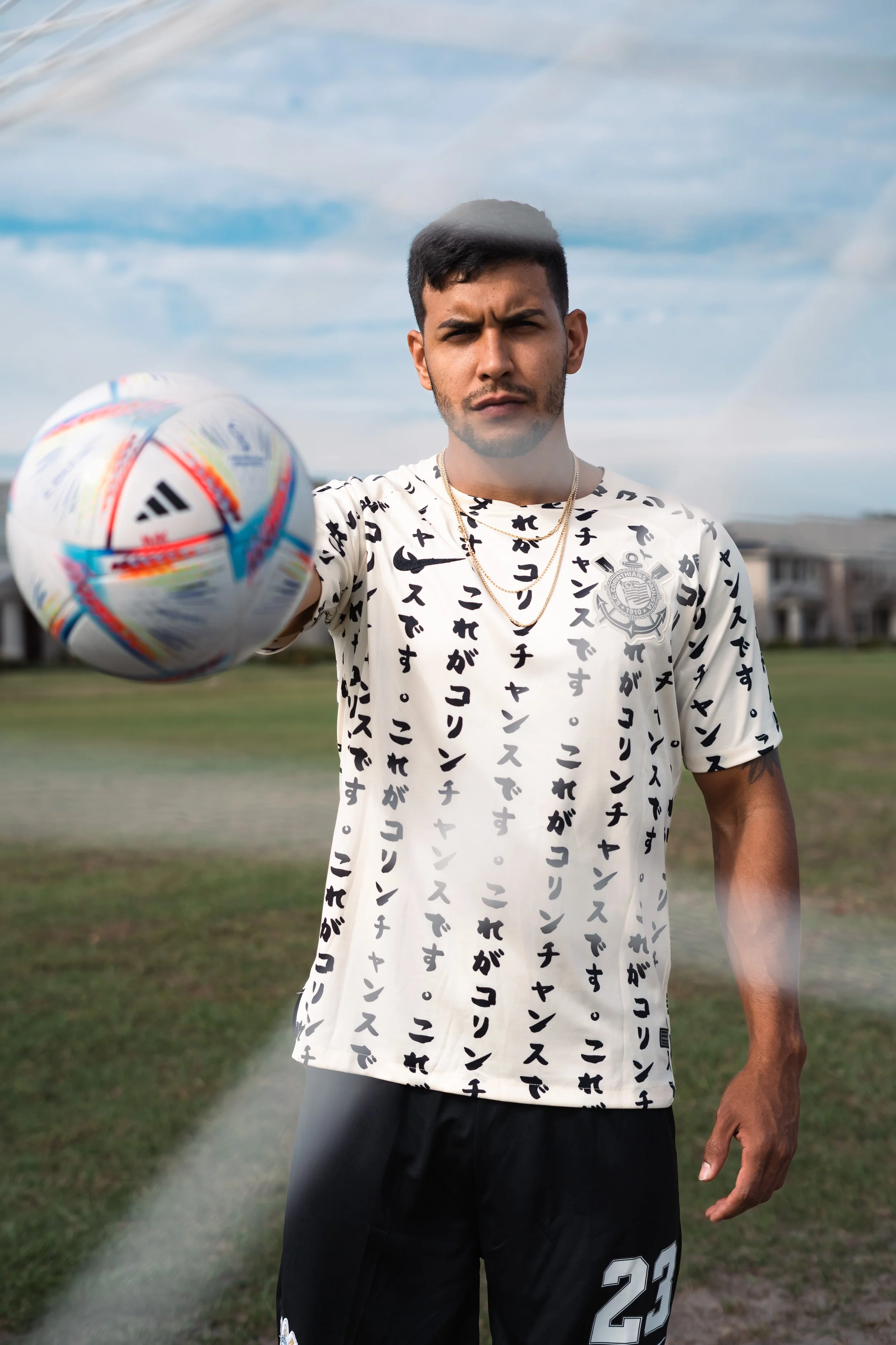 A young man holding a soccer ball on a grassy field, with a serious expression and a cloudy sky in the background.