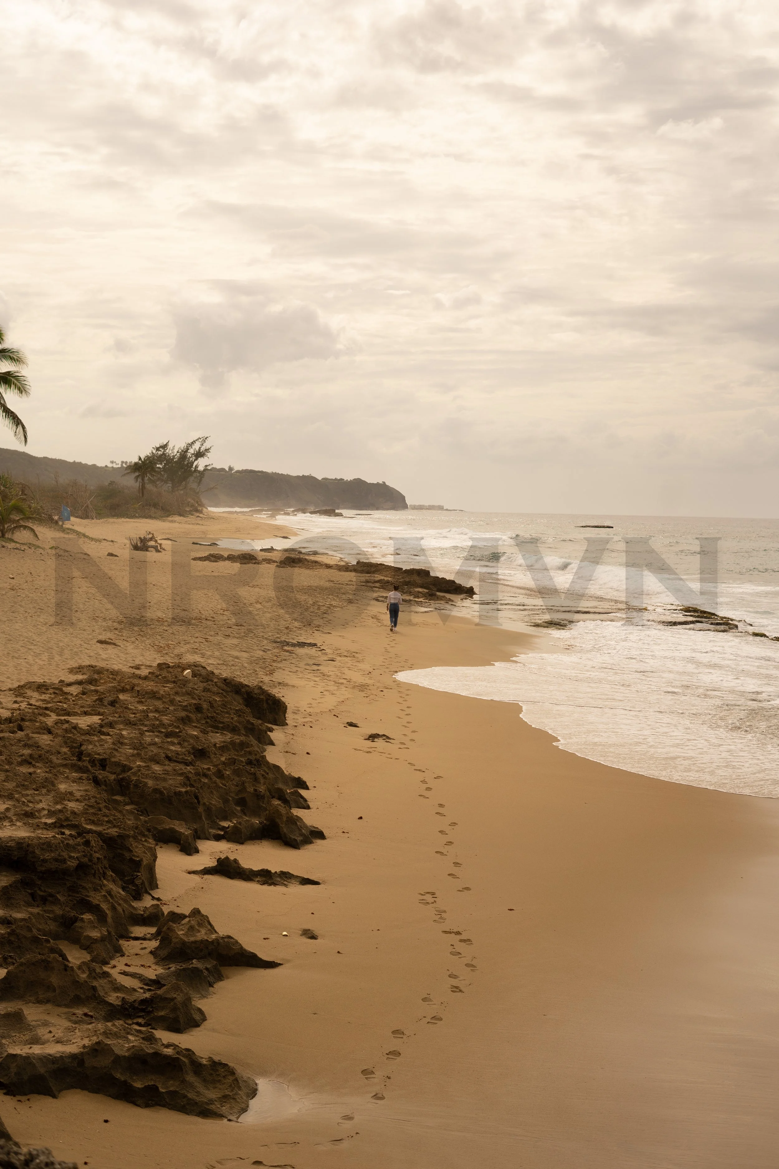 A person walking along a sandy beach with rocks, footprints, a few palm trees, and distant cliffs under cloudy skies.