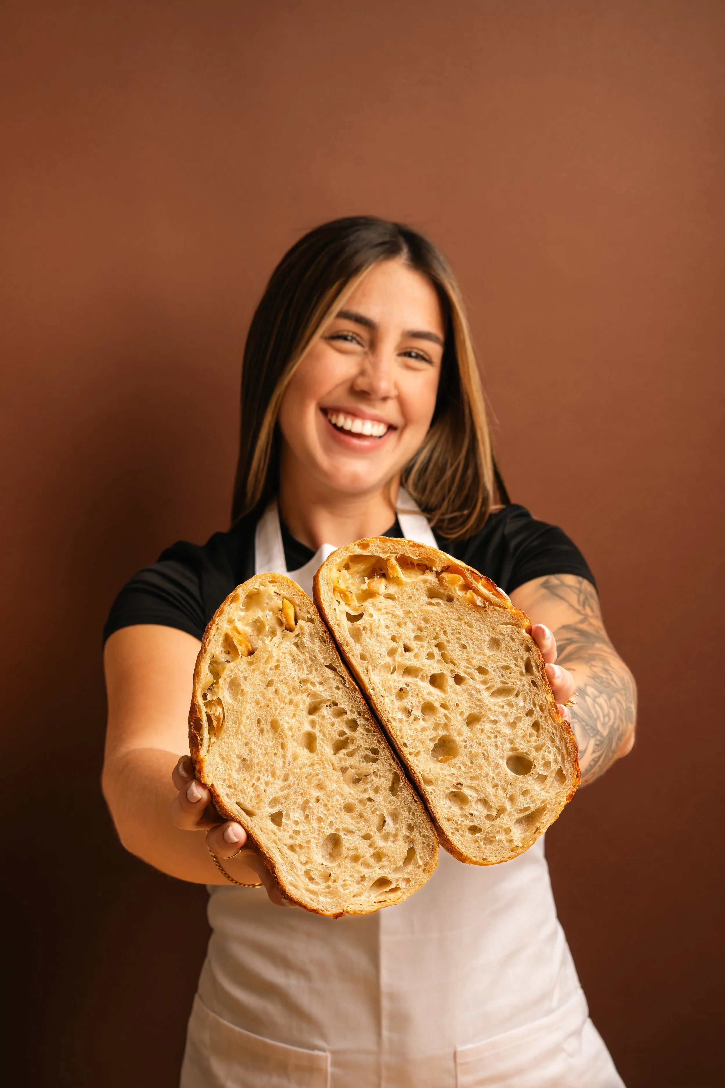 Woman with tattoos on arms holding a large sliced loaf of bread, smiling, against a brown background.