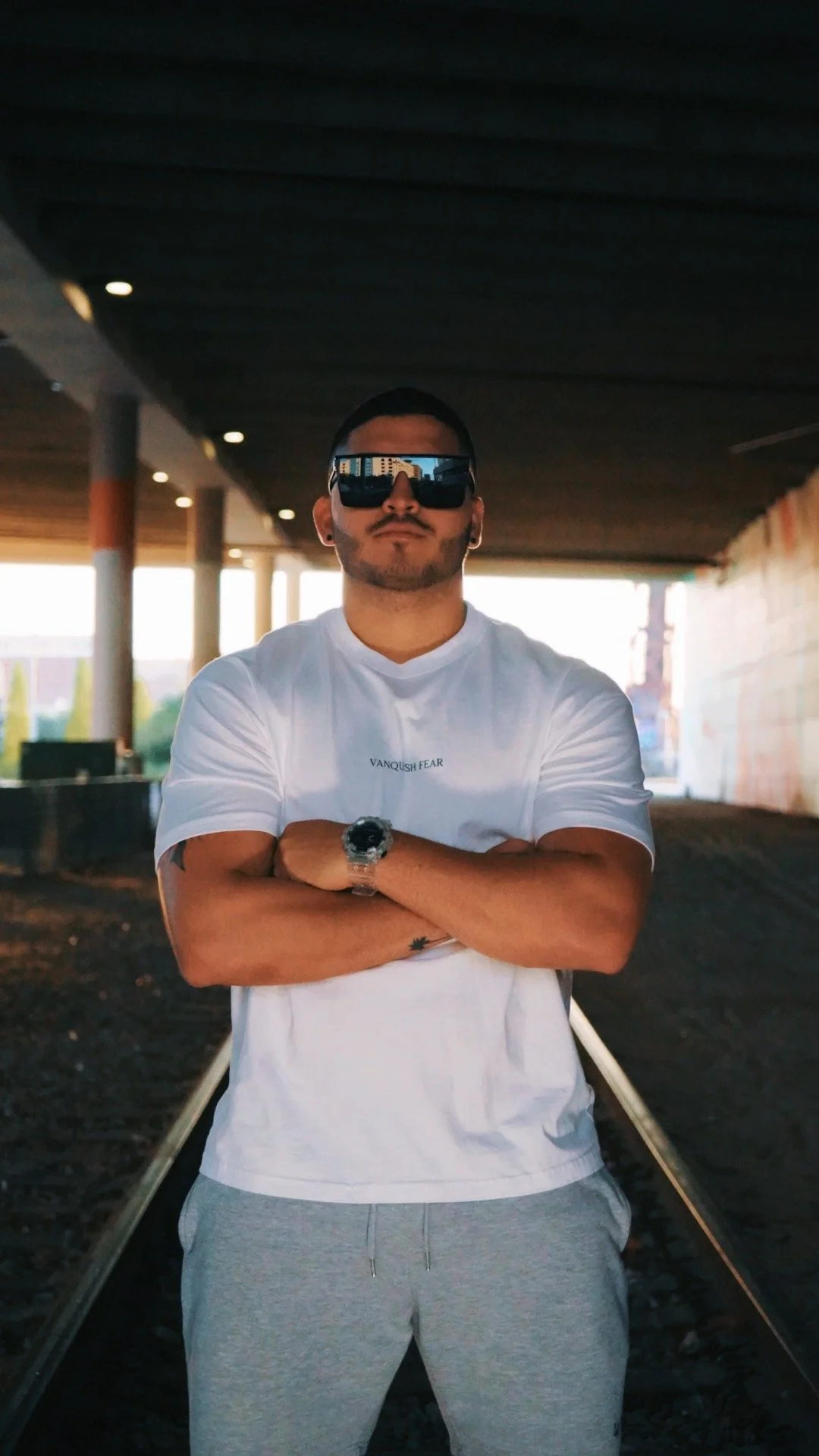 A man with short dark hair, sunglasses, and a beard stands with crossed arms under a bridge or tunnel at sunset.
