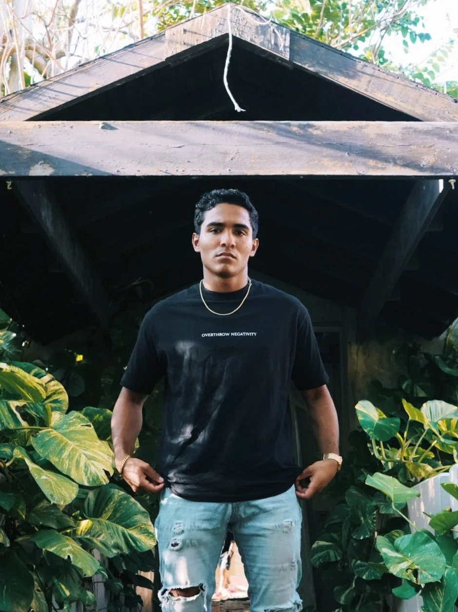 A young man standing outdoors in front of a small wooden house, surrounded by lush green plants. He is wearing a black t-shirt with white text, distressed jeans, a gold necklace, and a watch.