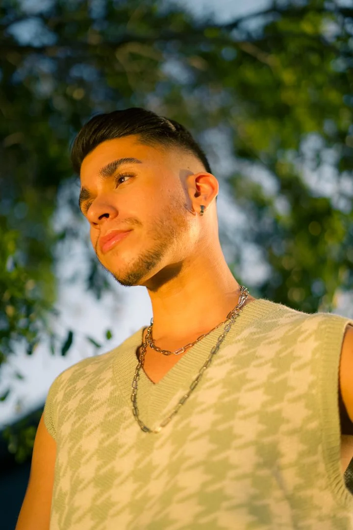 A young man with short dark hair and a trimmed beard, wearing earrings, layered necklaces, and a sleeveless sweater with a houndstooth pattern, posed outdoors with trees in the background.