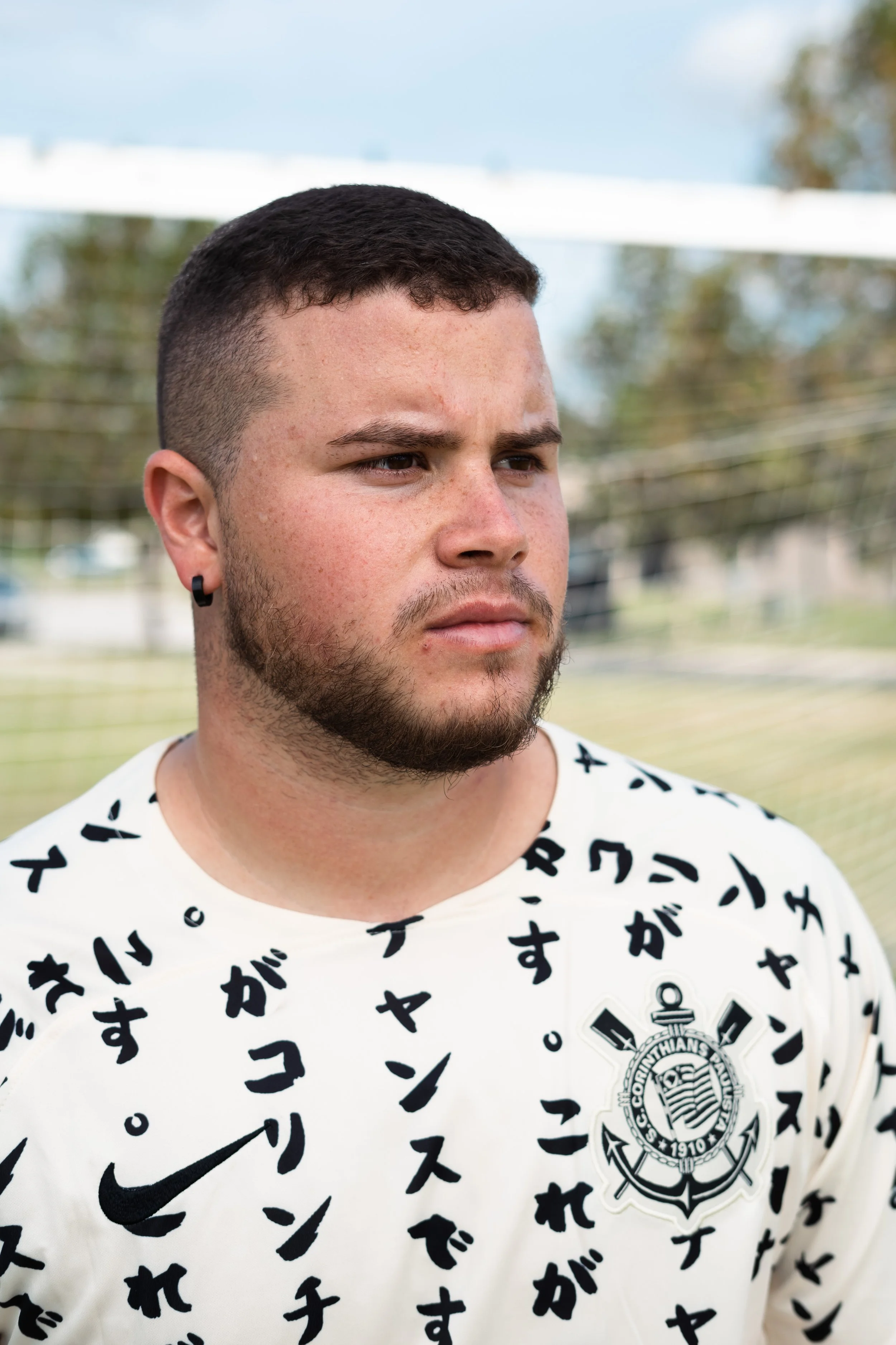 Close-up of a young man with short dark hair, a beard, and earrings, wearing a white sports jersey with black Japanese characters and a logo of SC Corinthians Paulista.