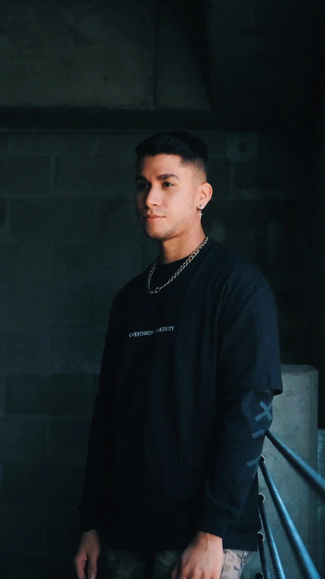 A young man with short dark hair, wearing a black T-shirt with white text and a black jacket, standing outdoors against a dark background with metal railings.