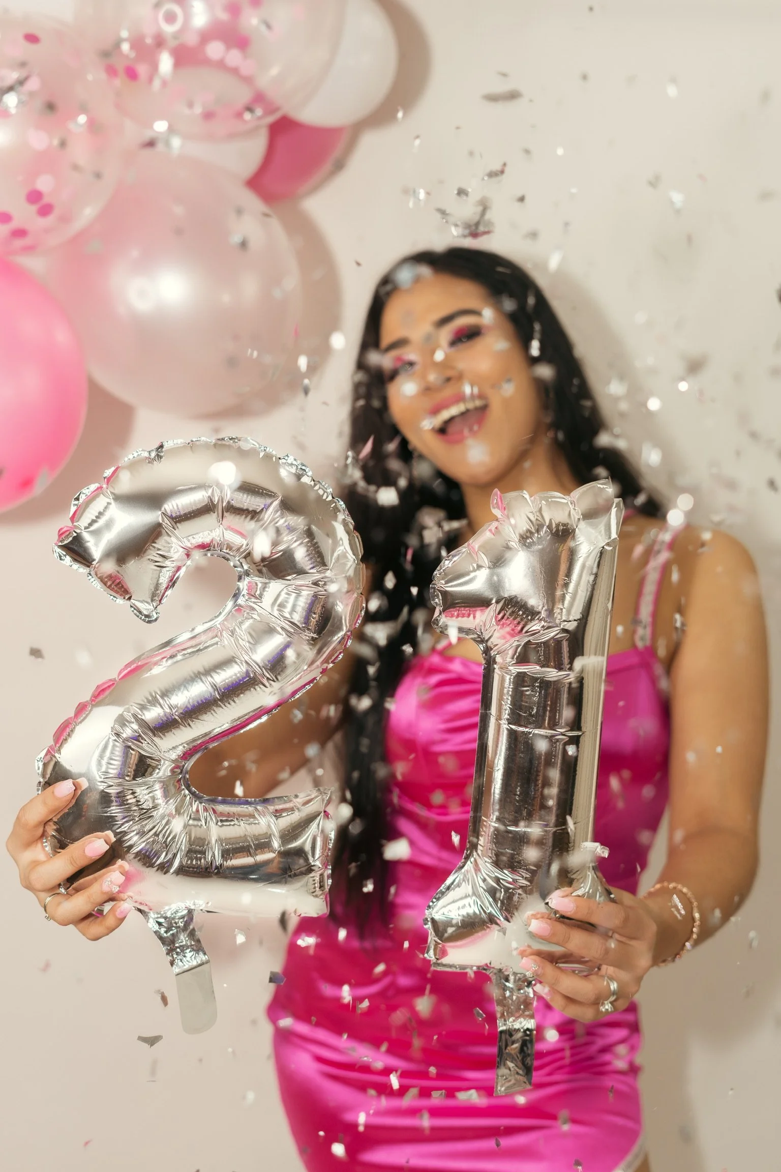 A woman wearing a pink dress celebrates New Year's Eve, holding balloons in the shape of the numbers 2 and 1, with confetti falling around her.