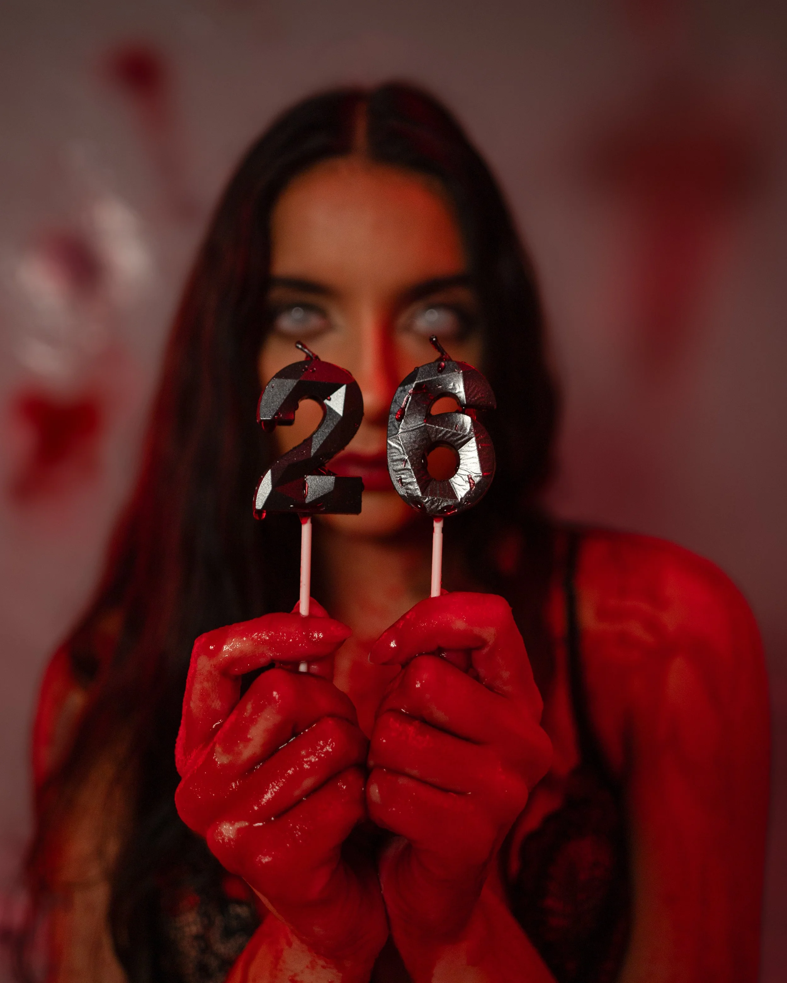A woman with long dark hair and pale eyes holds two candles shaped like the numbers 2 and 6, with her hands covered in red liquid resembling blood, in a dimly lit space with a red hue.