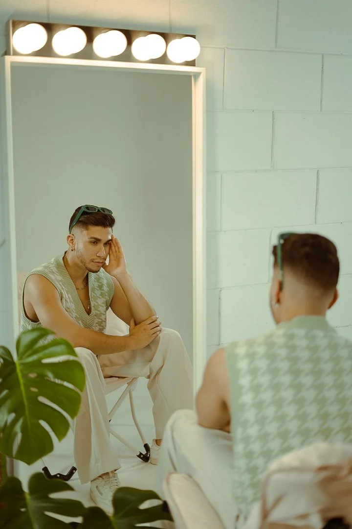A man sitting in front of a mirror, looking at himself with a contemplative expression. He is wearing sunglasses on his head, a sleeveless sweater, and white pants, with a plant partially visible in the foreground.