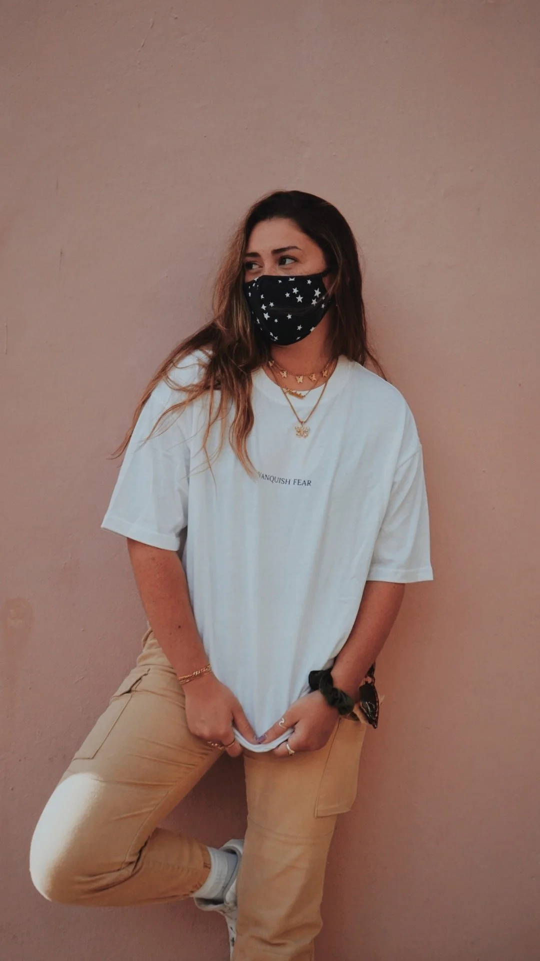 A young woman with wavy brown hair wearing a black face mask with stars, a white oversized t-shirt, tan pants, white sneakers, and black wrist accessories, standing against a peach-colored wall.