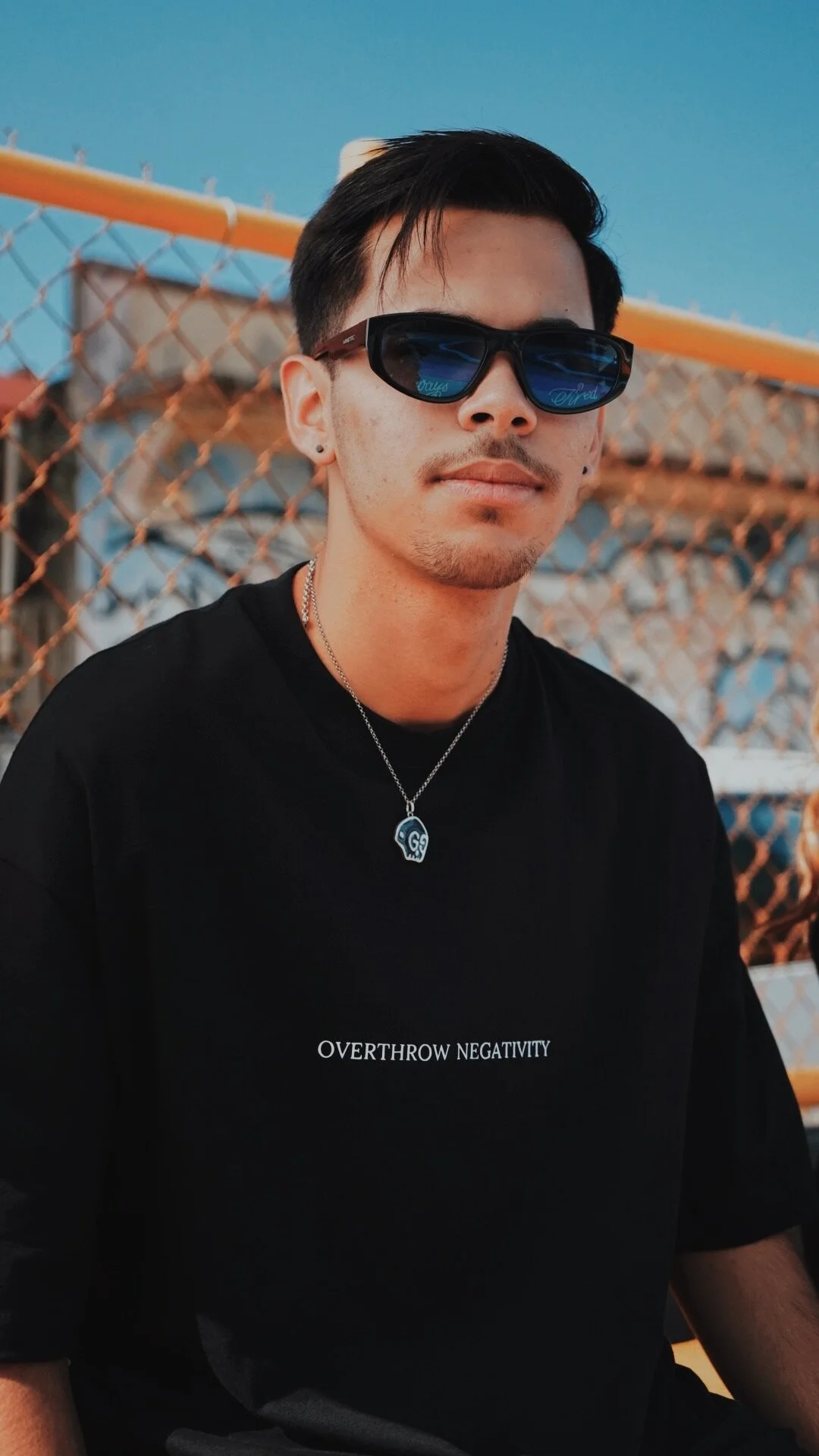 A young man with black hair, sunglasses, and a black T-shirt that says 'OVERTROW NEGATIVITY,' wearing a necklace with a skull pendant, sits outdoors near a chain-link fence with a building in the background.