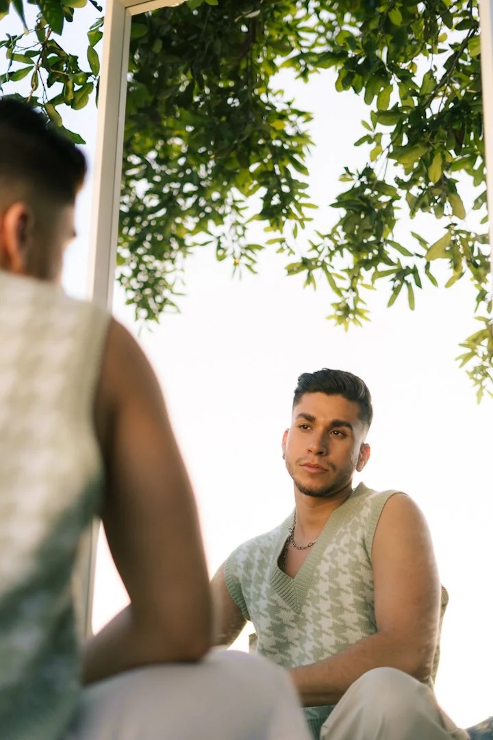 Two men sitting outdoors near a mirror, with lush green foliage overhead and sunlight filtering through the trees.