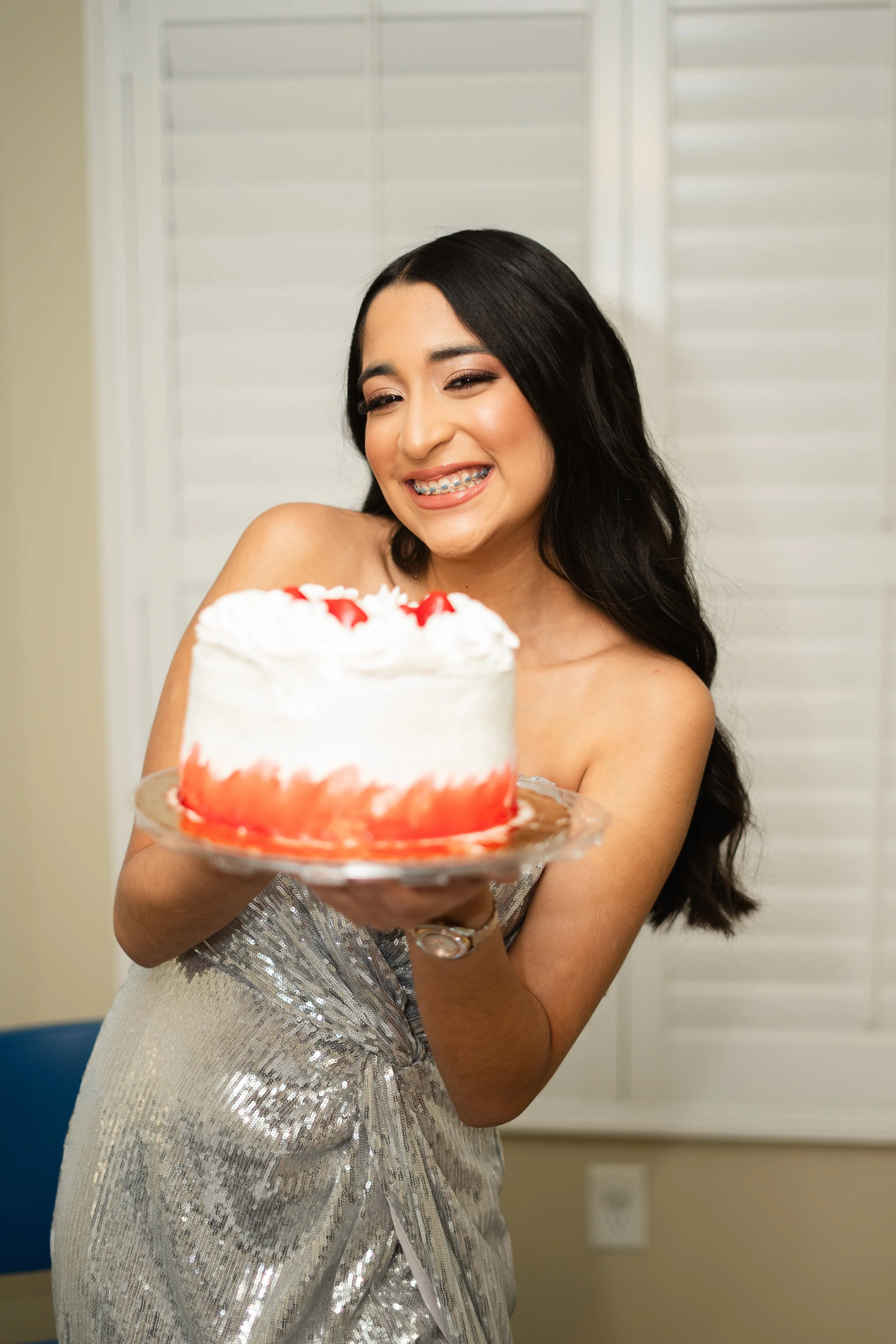 A woman with long dark hair and braces holding a white frosted cake decorated with red sauce or berries, smiling in a room with white blinds.