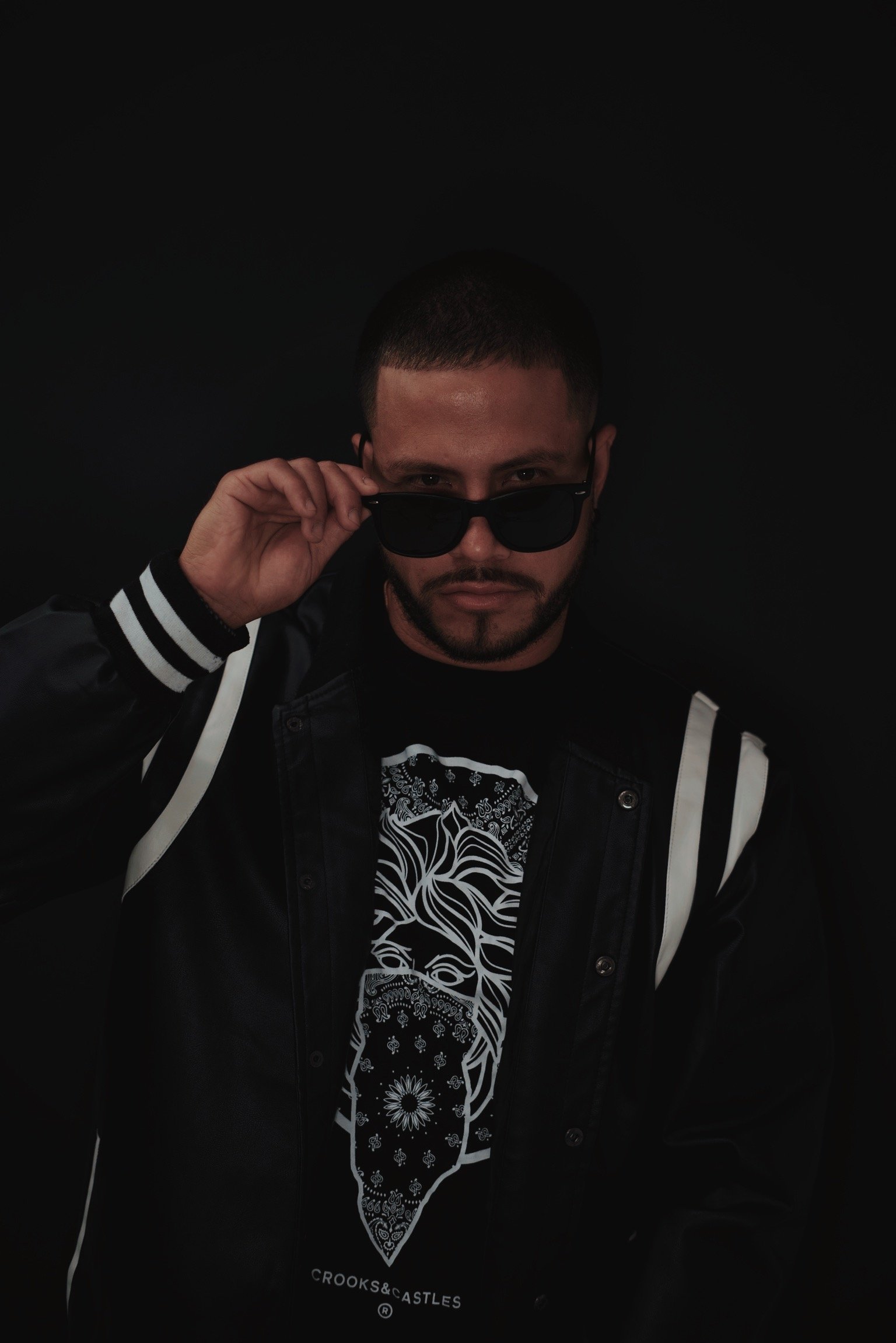 A man wearing sunglasses and a black jacket, lifting his sunglasses with his right hand, against a black background.