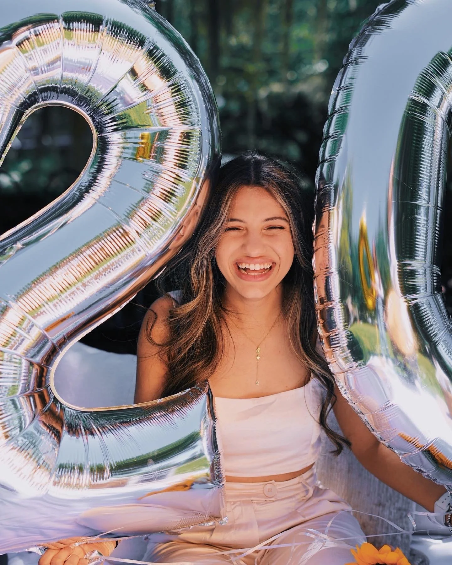 A young woman smiling and laughing outdoors, holding large silver foil balloons shaped as the numbers 2 and 0, in a park or garden setting.