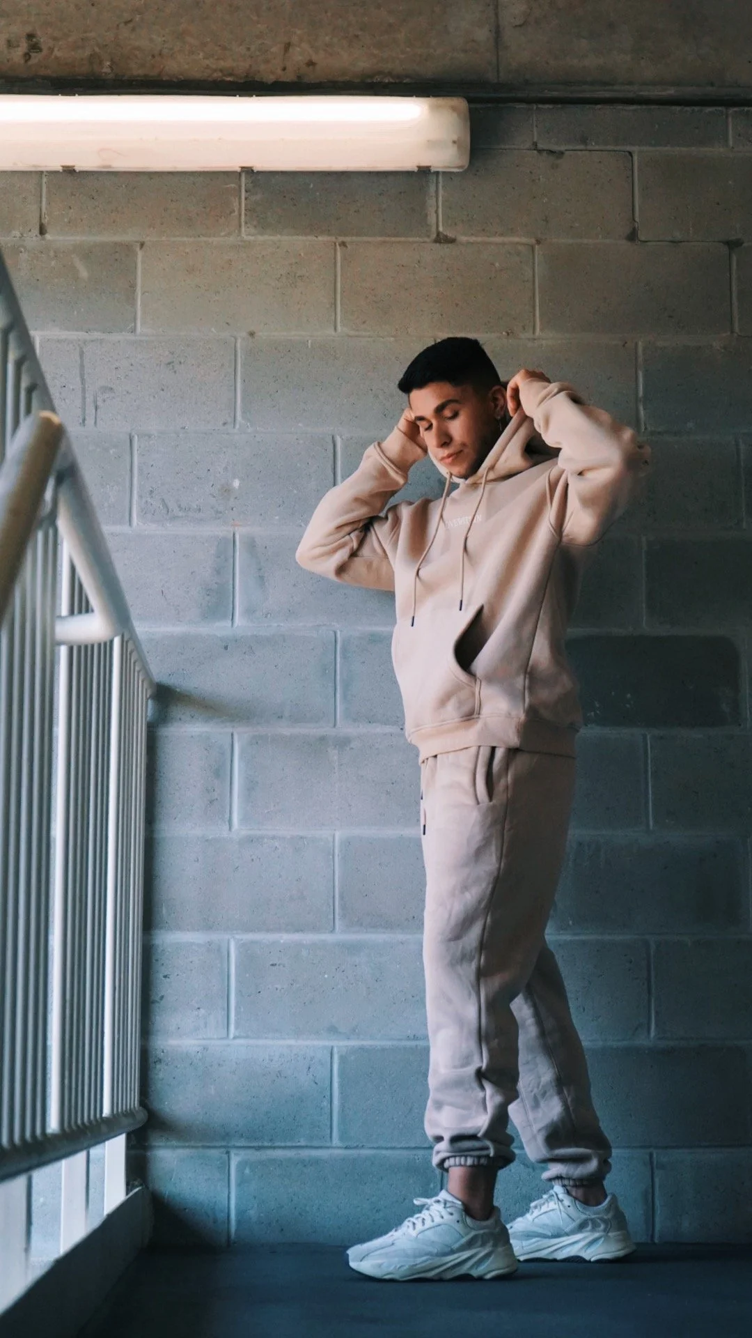 A young man in light beige hoodie and matching joggers standing against a concrete block wall, adjusting his hoodie with eyes closed and a relaxed expression.