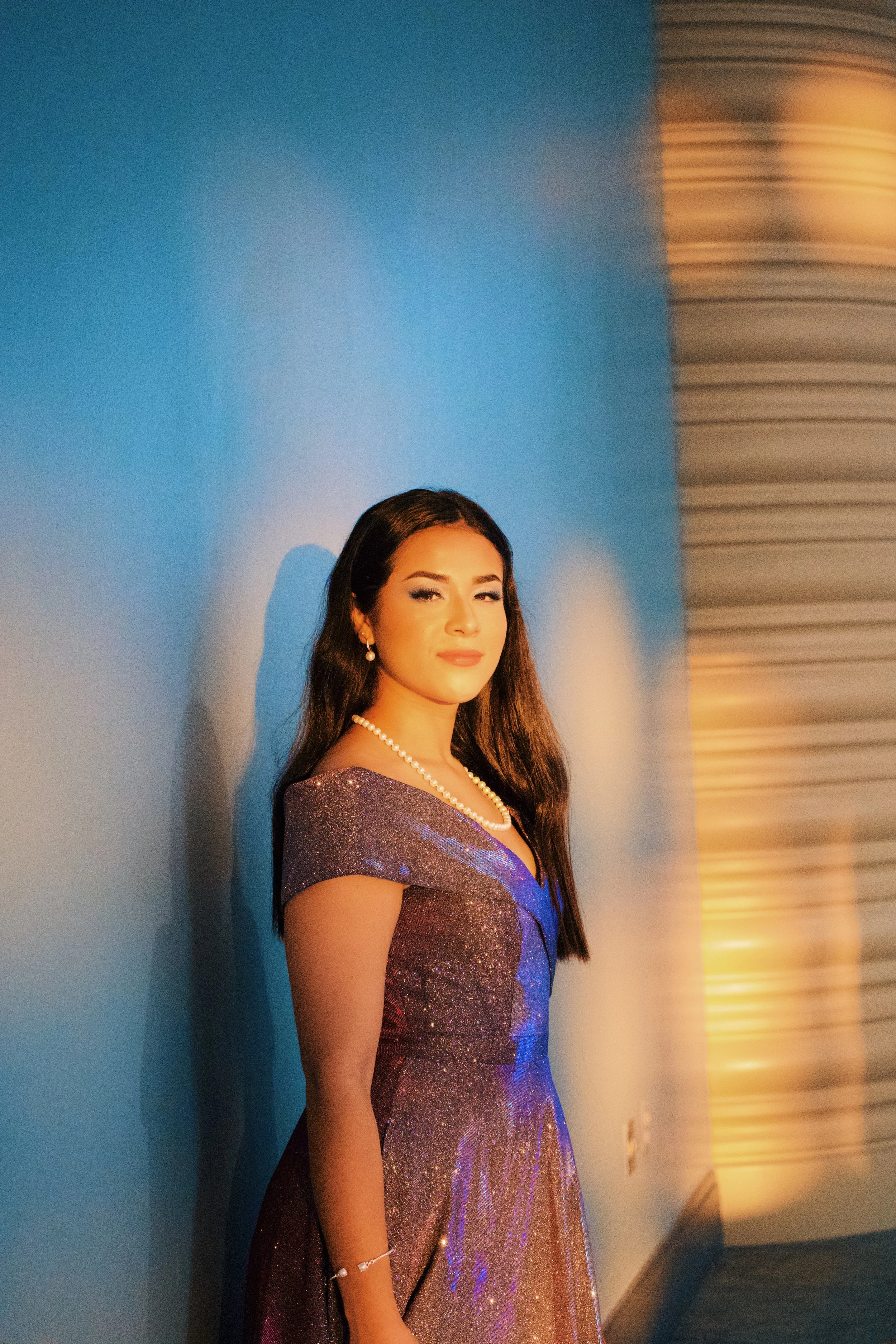 A woman in a shimmering purple dress stands against a blue wall with a shadow cast behind her. She is wearing pearl jewelry, including a necklace, earrings, and a bracelet, and has long dark hair styled down. She appears to be in an elegant setting w