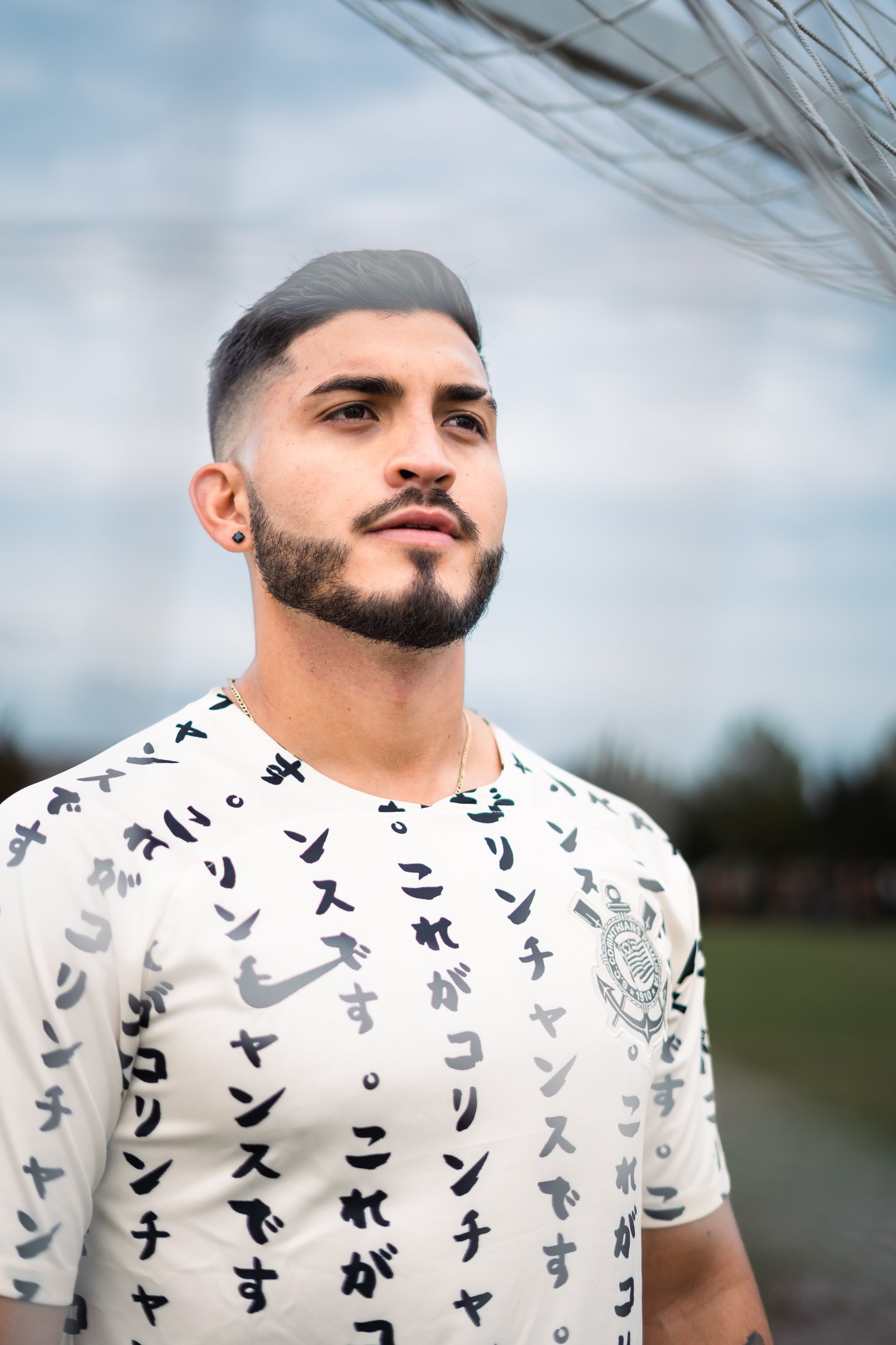 A young man with dark hair, beard, and earrings wearing a white sports jersey with Japanese characters and a crest, standing outdoors on a cloudy day.