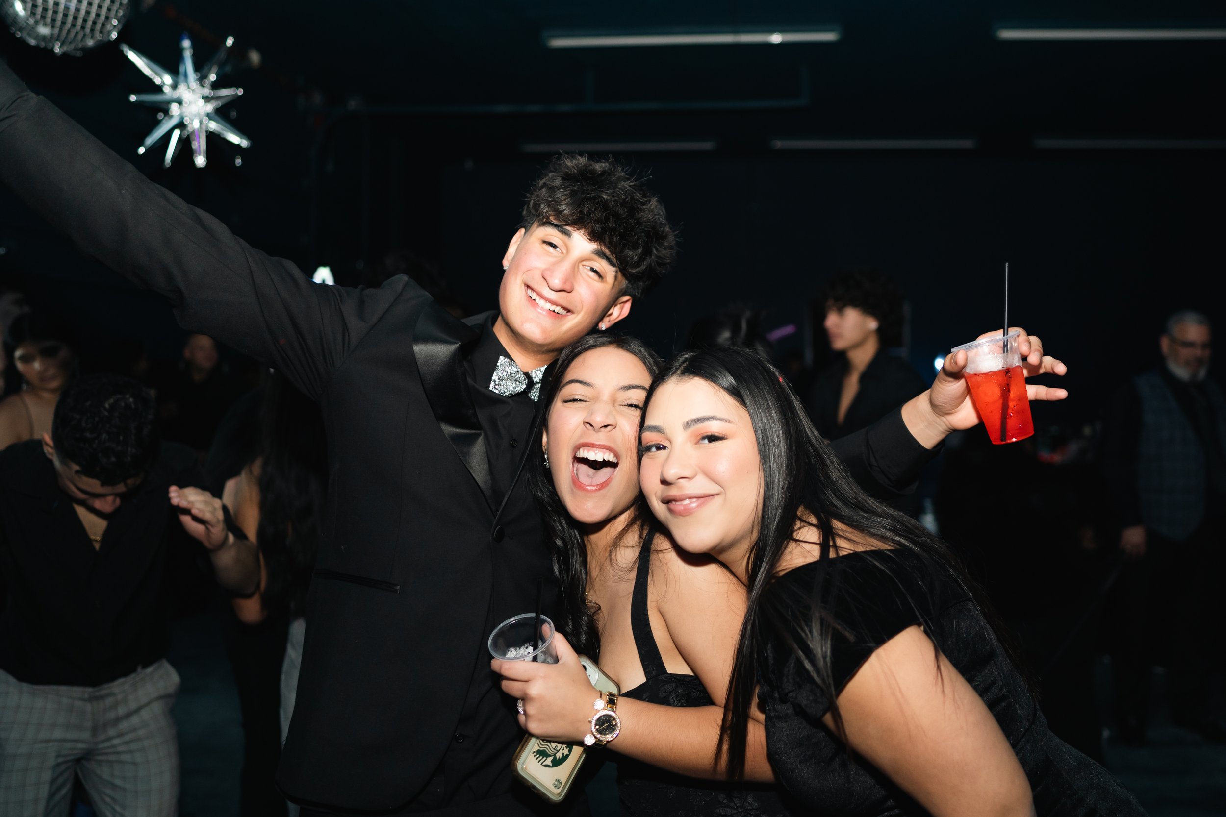 Three young adults at a party, smiling and holding drinks, with other people in the background.