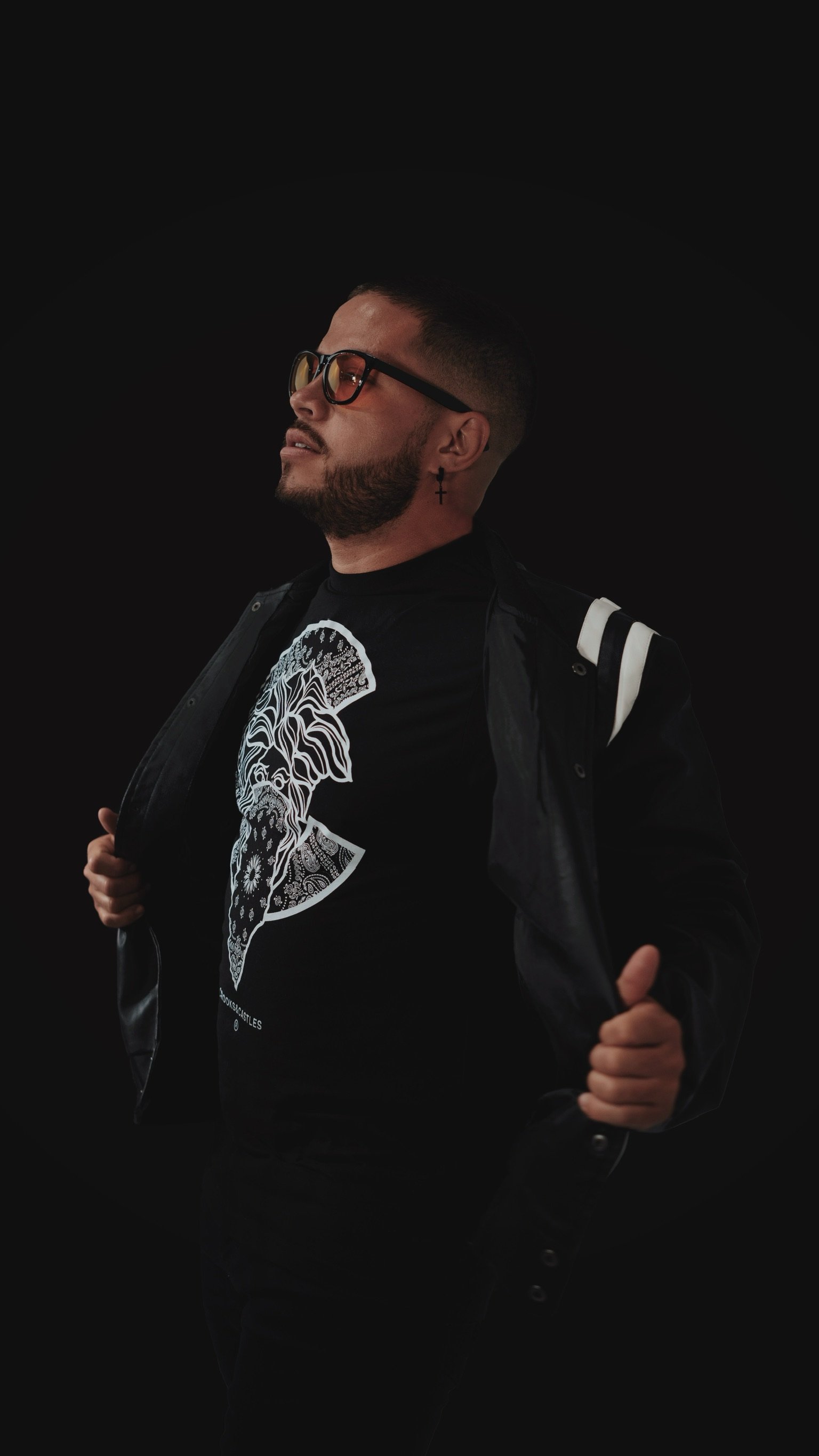 A man wearing glasses, a black T-shirt with a white graphic, and a black jacket, standing against a black background.