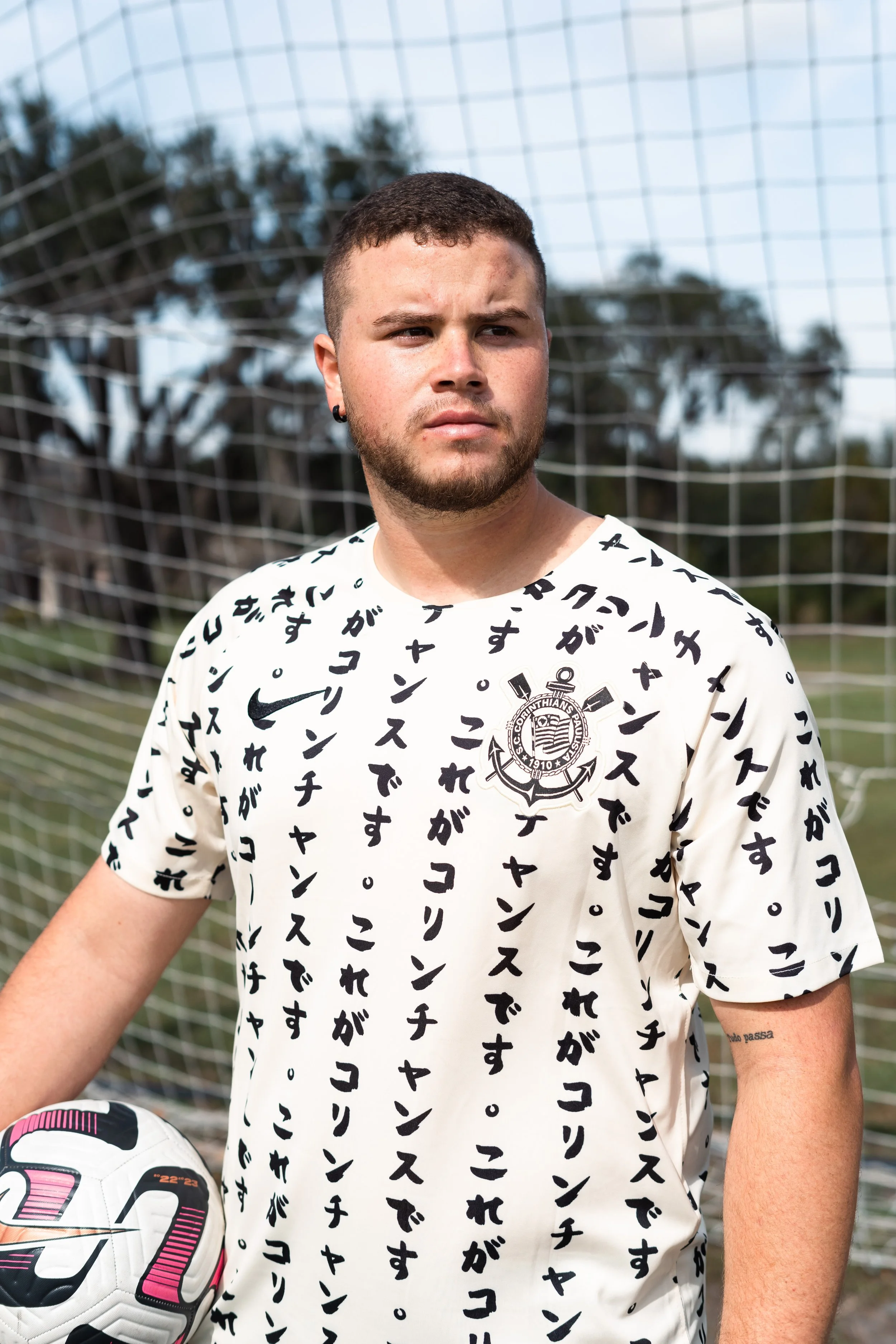 Young man in a white soccer jersey with Japanese characters and a club logo, standing on a soccer field with goal netting in the background, holding a soccer ball.
