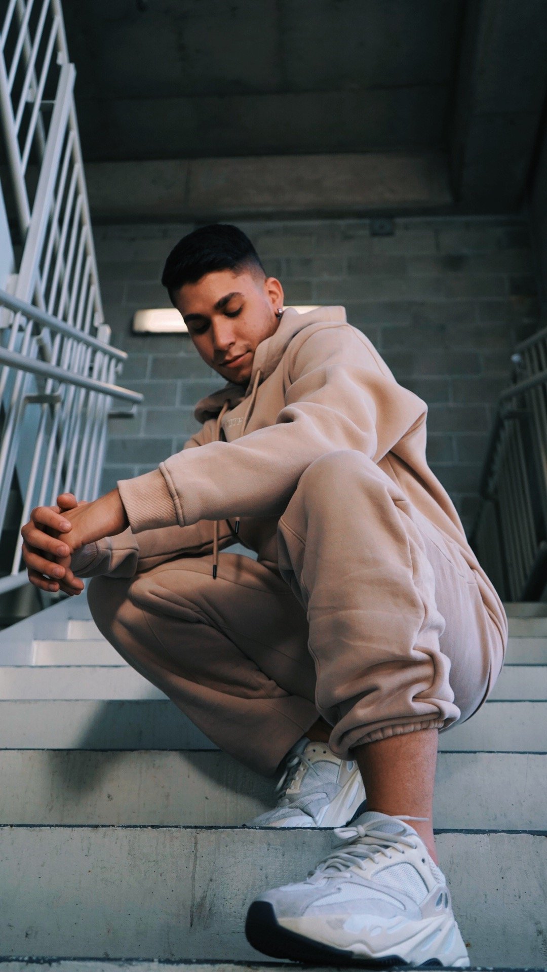 A young person in beige hoodie and sweatpants squatting on outdoor stairs, looking down with hands clasped.