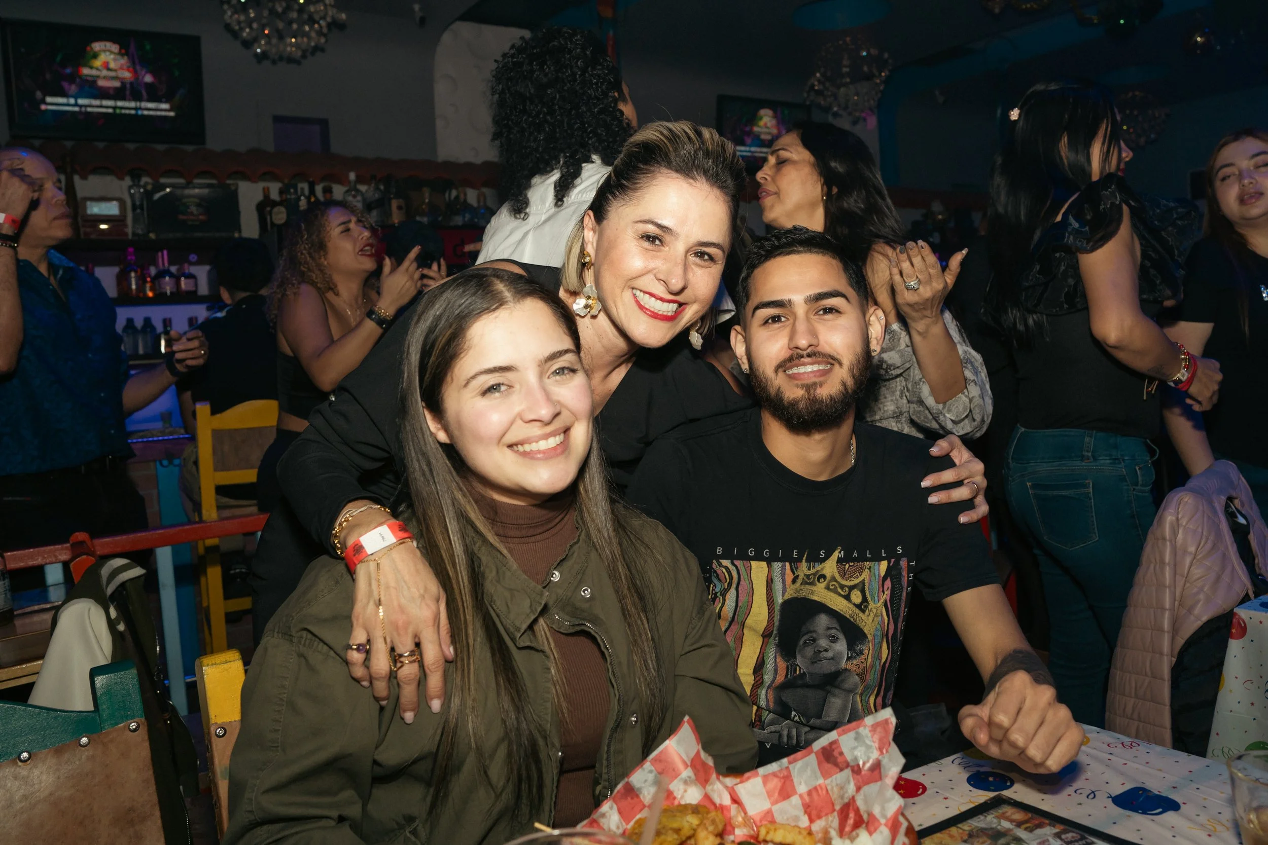 Three people smiling and sitting at a table in a lively party or celebration. The woman in the middle is standing behind the other two, with her arms around them. The background shows other people dancing and enjoying the event.