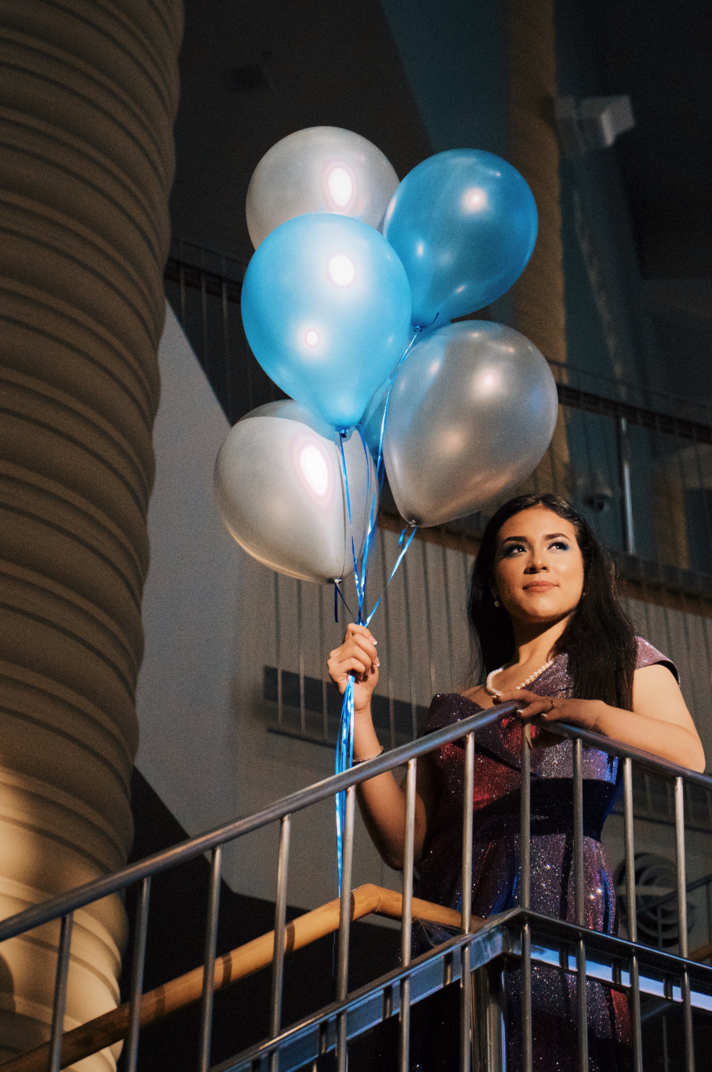 A young woman with dark hair wearing an elegant dress holding a bunch of blue and silver balloons on a balcony at night.