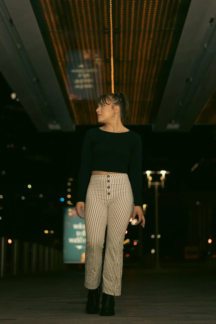 A woman standing under a modern wooden ceiling at night, wearing a black long-sleeve crop top, striped high-waisted pants, and black boots.