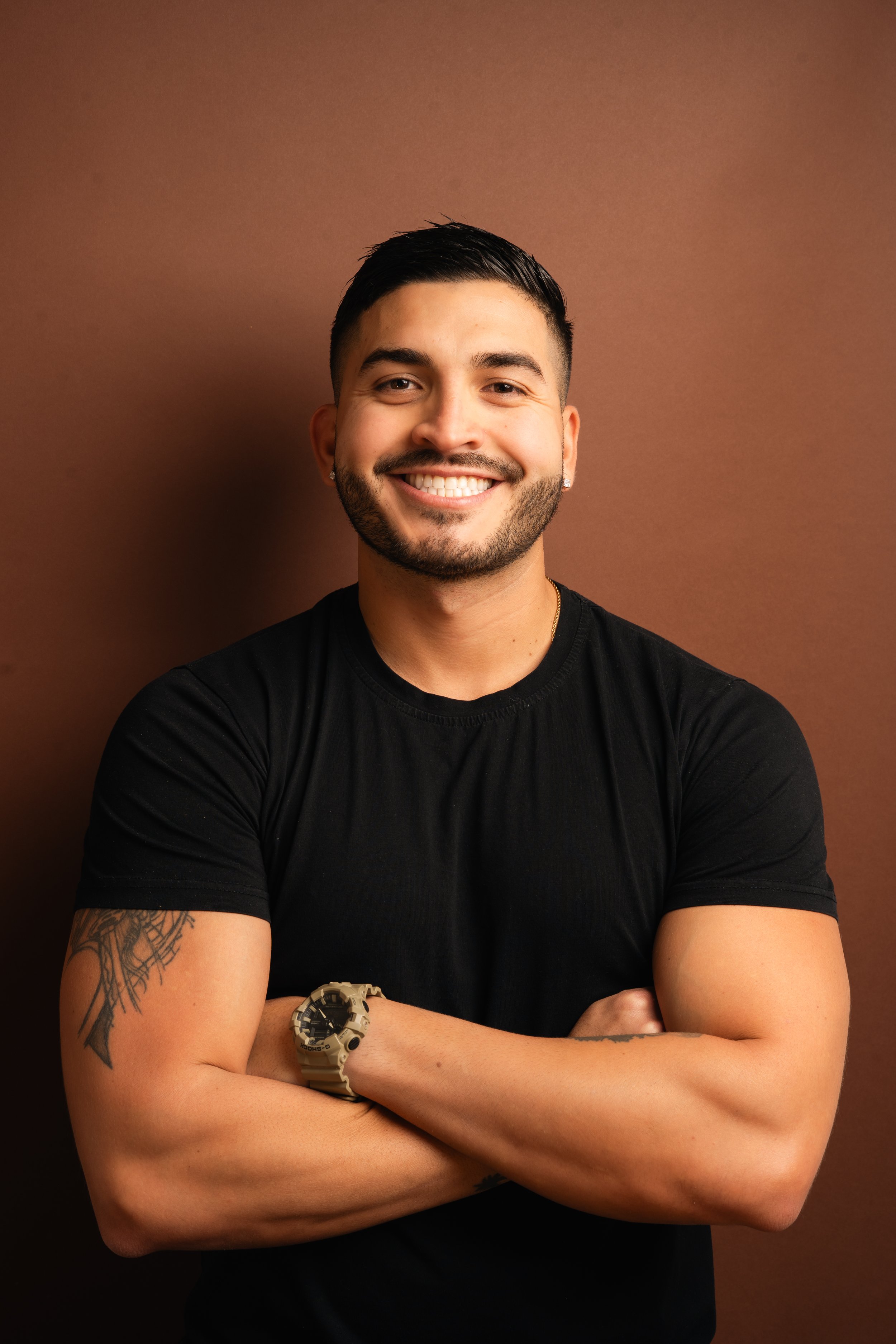 A young man with short black hair and a beard, smiling with arms crossed, wearing a black t-shirt, a watch, and earrings, standing against a brown background.