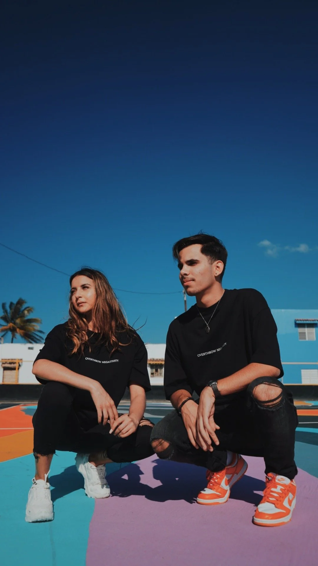 Two young people, a woman and a man, squatting on a colorful painted playground under a clear blue sky, wearing black T-shirts and ripped jeans.